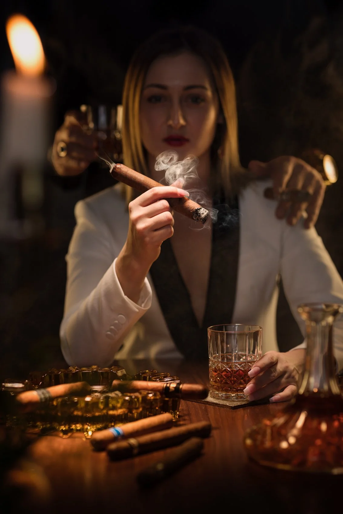 A woman in a white blazer holding a cigar and a glass of drink, sitting at a bar surrounded by cigars and a candle.