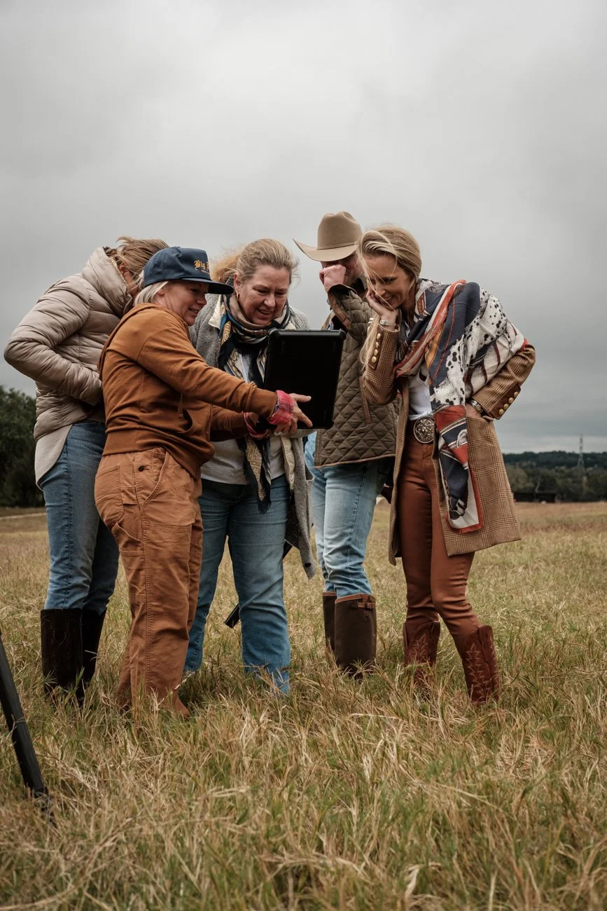 Five women dressed in outdoor clothing stand in a grassy field, gathered around a laptop, looking at the screen and smiling, under a cloudy sky.