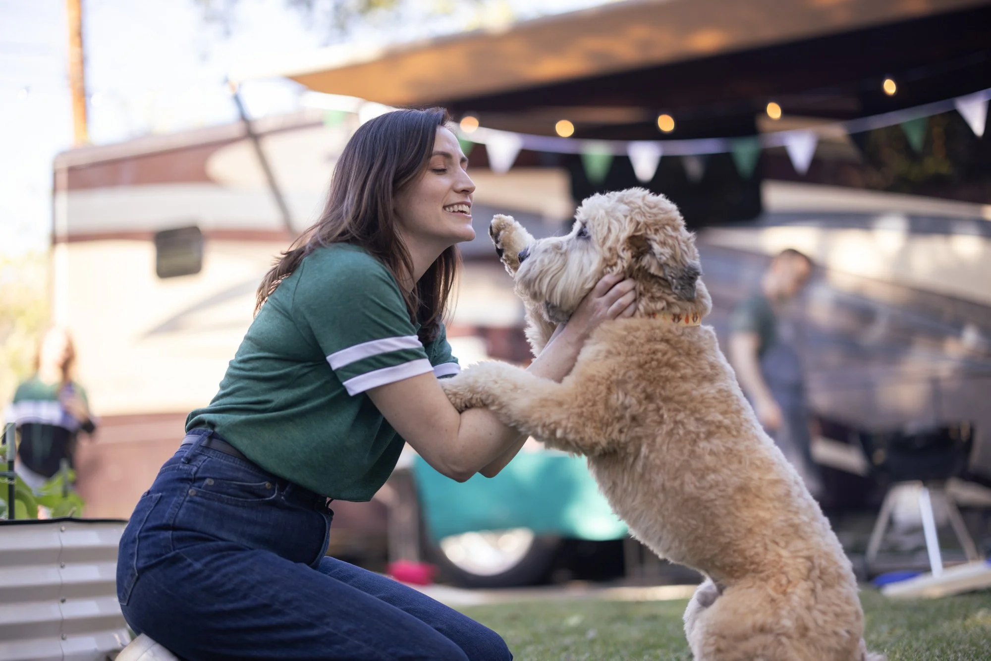 A woman with long dark hair in a green shirt and blue jeans smiles as she plays with a large, fluffy, light-colored dog at an outdoor event under a canopy decorated with triangular flags and string lights.