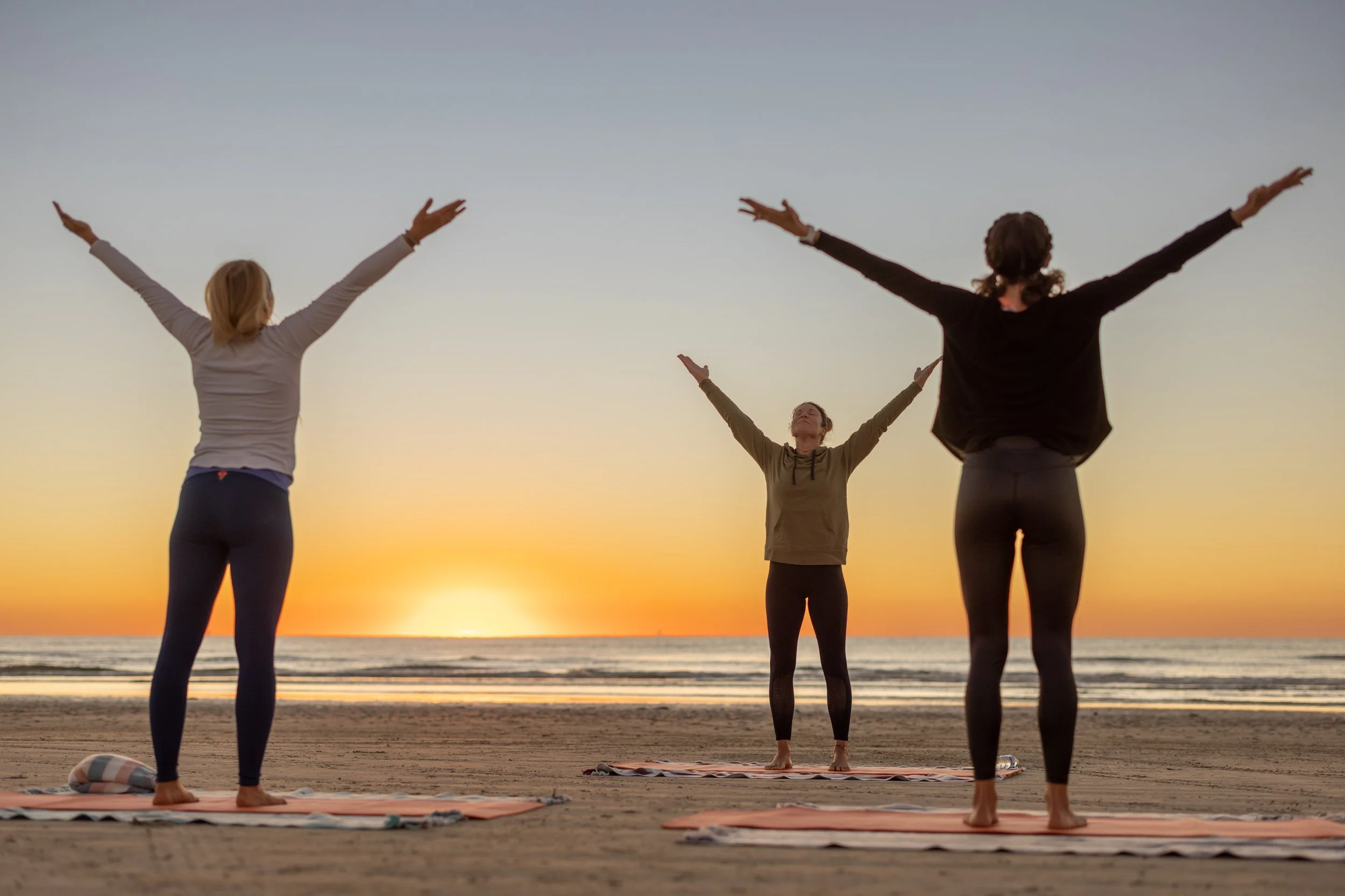 People practicing yoga on a beach during sunset with arms raised in a greeting pose.