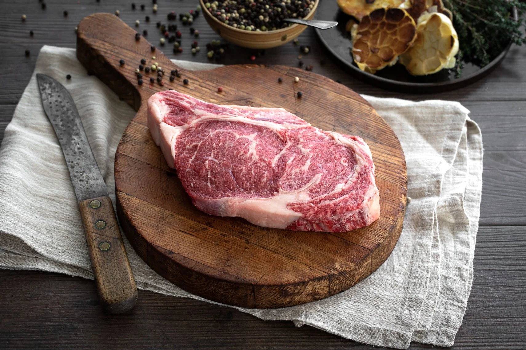 A raw, marbled beef steak on a wooden cutting board with a knife nearby. In the background, there are bowls of peppercorns and garlic, and some herbs on a dark wooden surface.