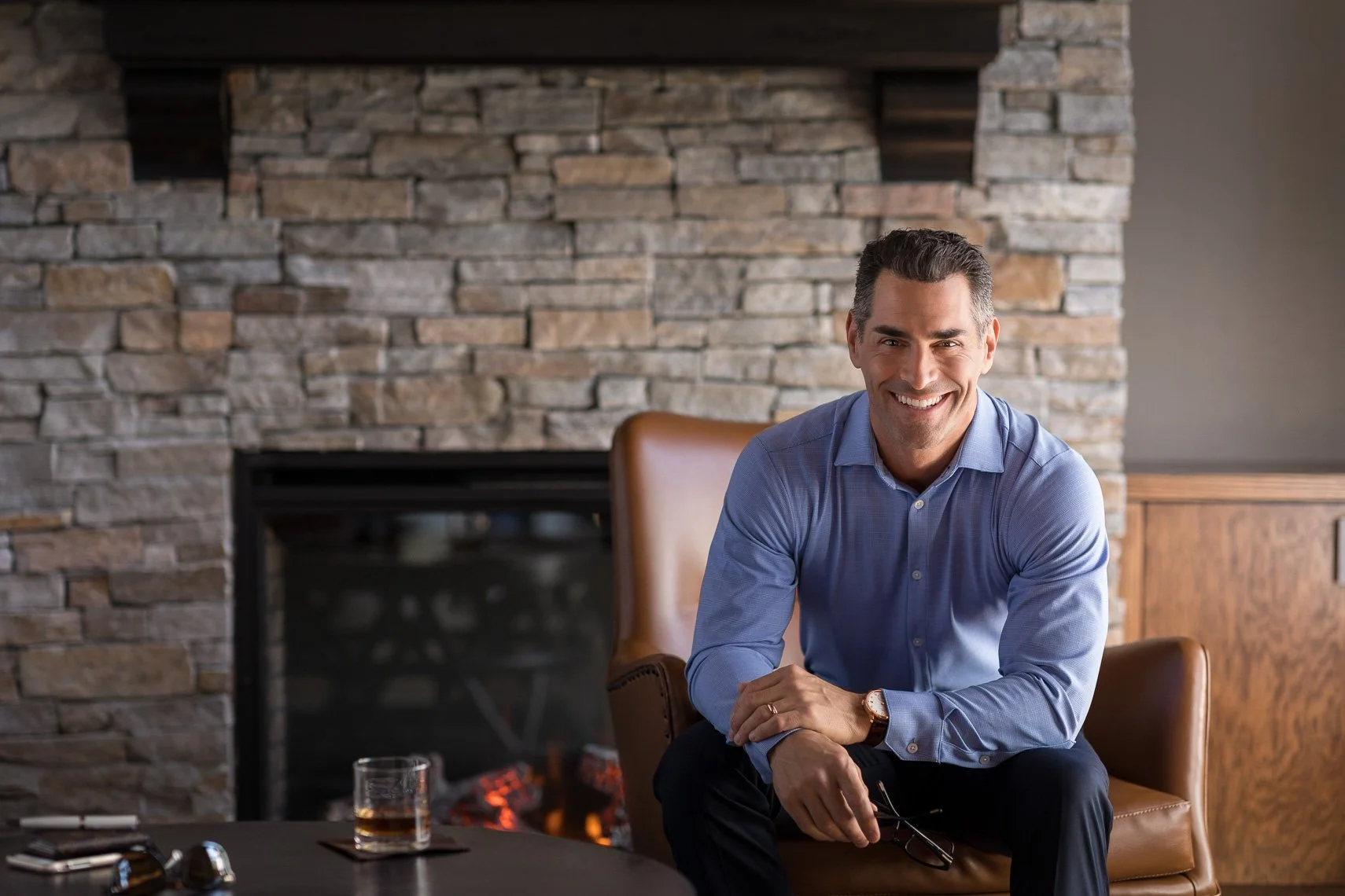 A smiling man in a blue dress shirt sitting on a brown leather chair in front of a stone fireplace with a glass of whiskey on a table nearby.