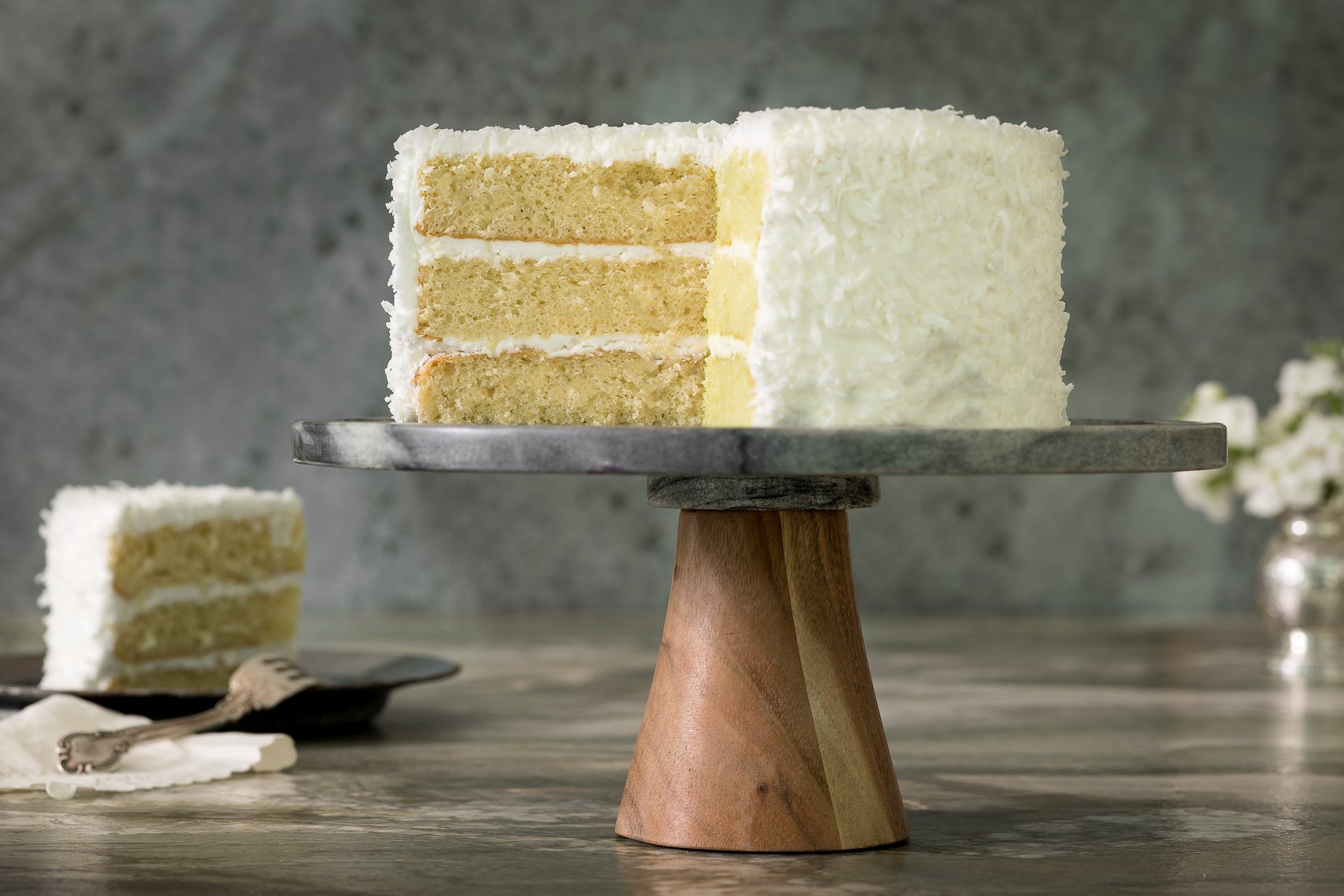 A slice of layered vanilla cake with white frosting on a cake stand, with a piece on a plate and a fork in the foreground.