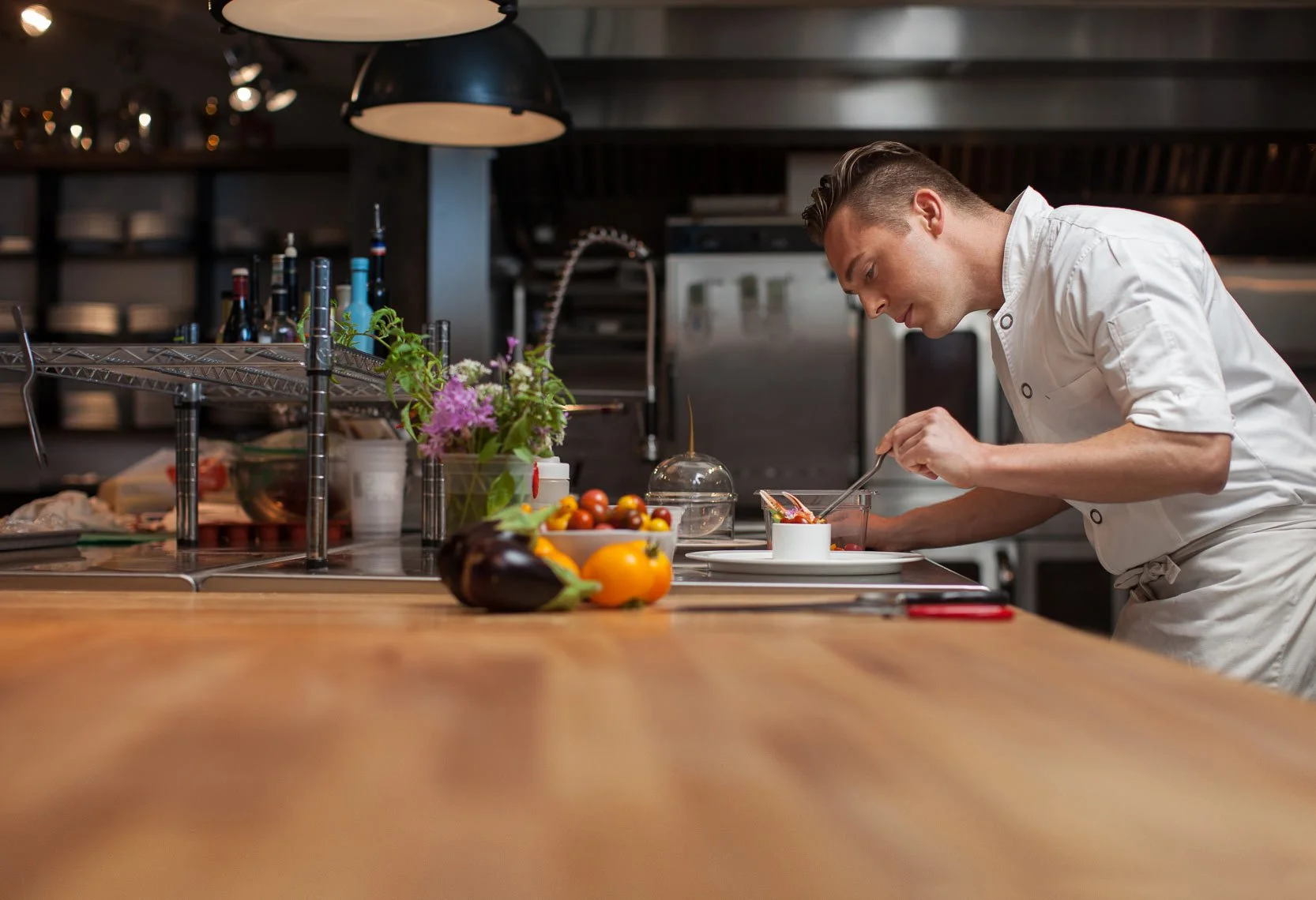 A chef in a white uniform preparing food in a modern kitchen with a cluttered counter, colorful vegetables, and a vase of flowers.