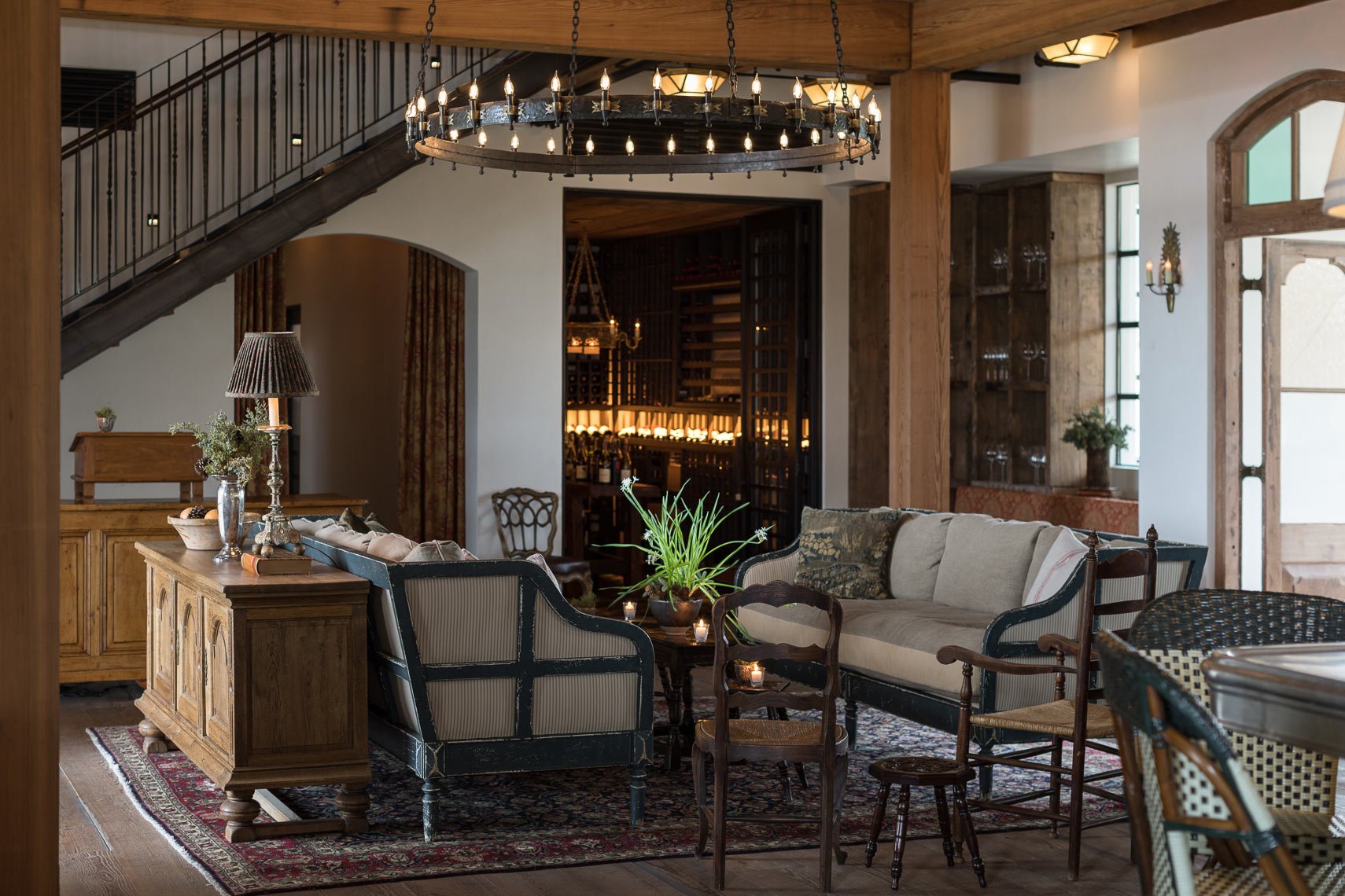 A cozy living room with a rustic style, featuring a large chandelier, a wooden sideboard with a lamp and plants, a cream-colored sofa with pillows, various wooden chairs, a potted plant on a coffee table, a patterned rug, and a wine cellar in the bac
