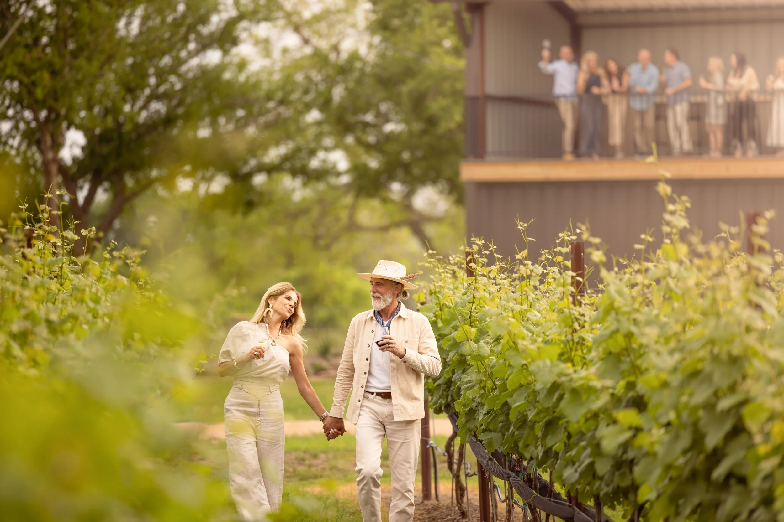 A couple walking through a vineyard holding hands, with a man wearing a hat and talking to a woman, while a group of people are observing from a balcony in the background.