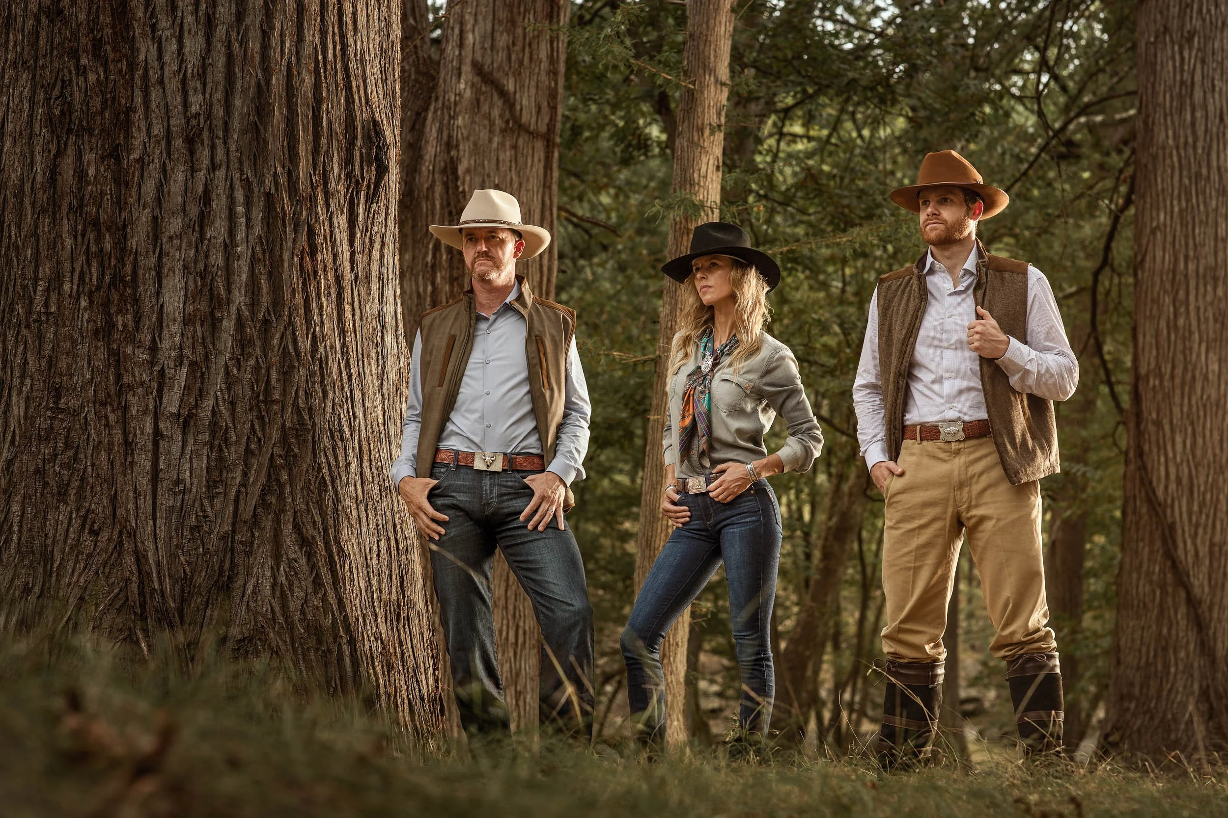 Three people standing among large trees in a forest, wearing cowboy hats and outdoor clothing, looking serious and contemplative.