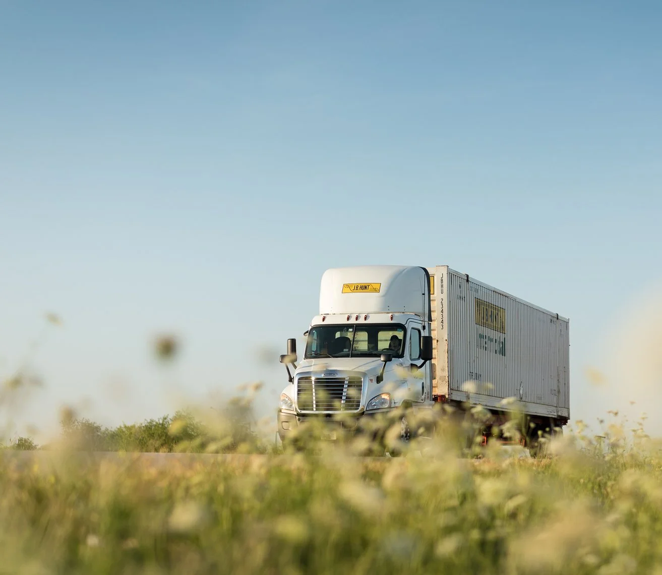 A white semi-truck driving on a rural road surrounded by yellow wildflowers, with a clear blue sky overhead.