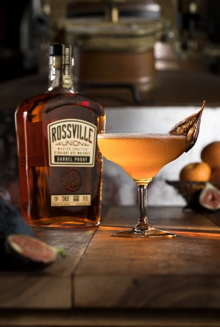 A glass of cocktail with a dried fruit garnish next to a bottle of Rossville Union whiskey on a wooden table. There are figs and oranges in the background.