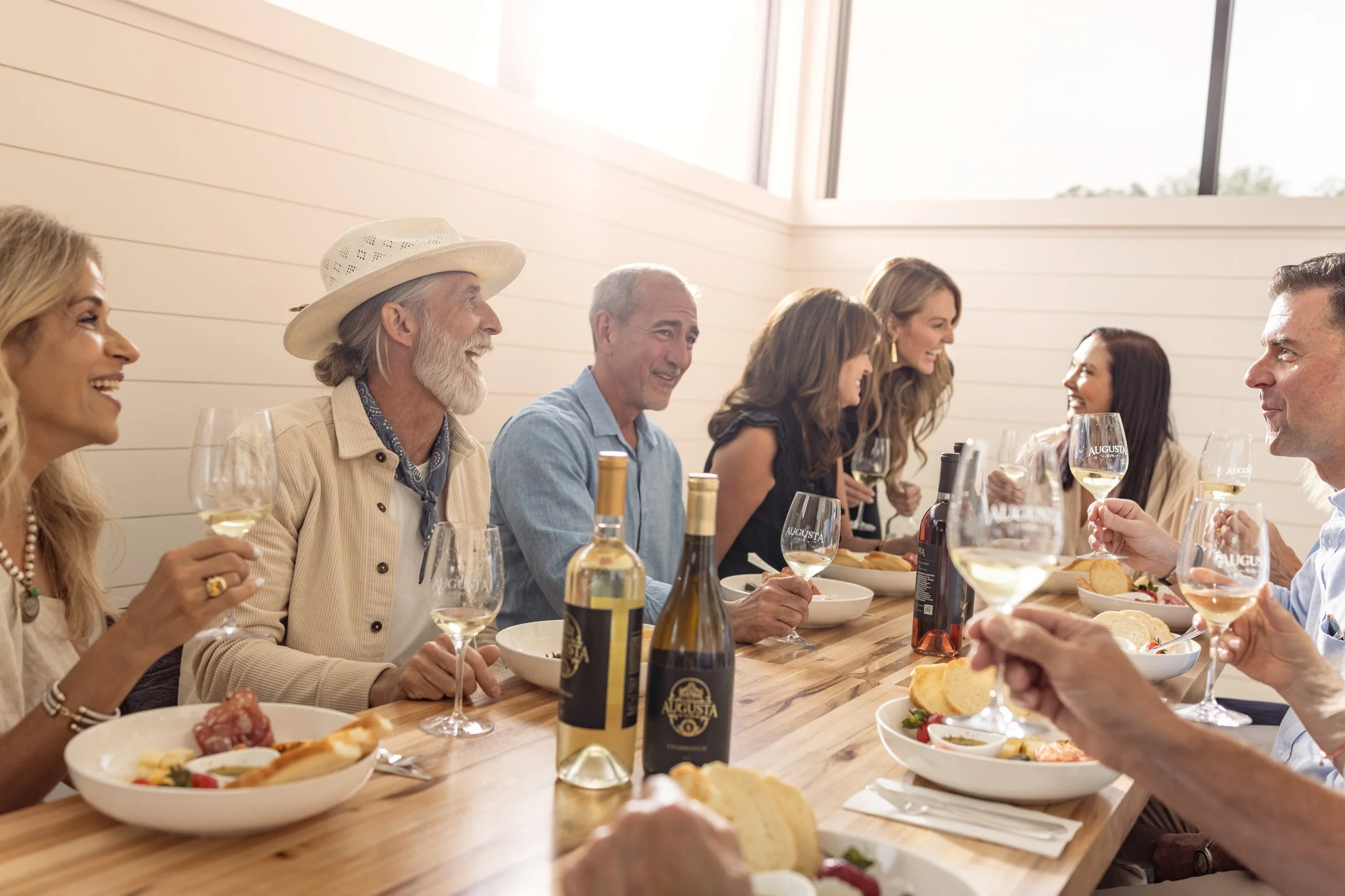 A group of people enjoying a meal at a wooden table with plates of food, wine glasses, and bottles, seated indoors with large windows letting in natural light.