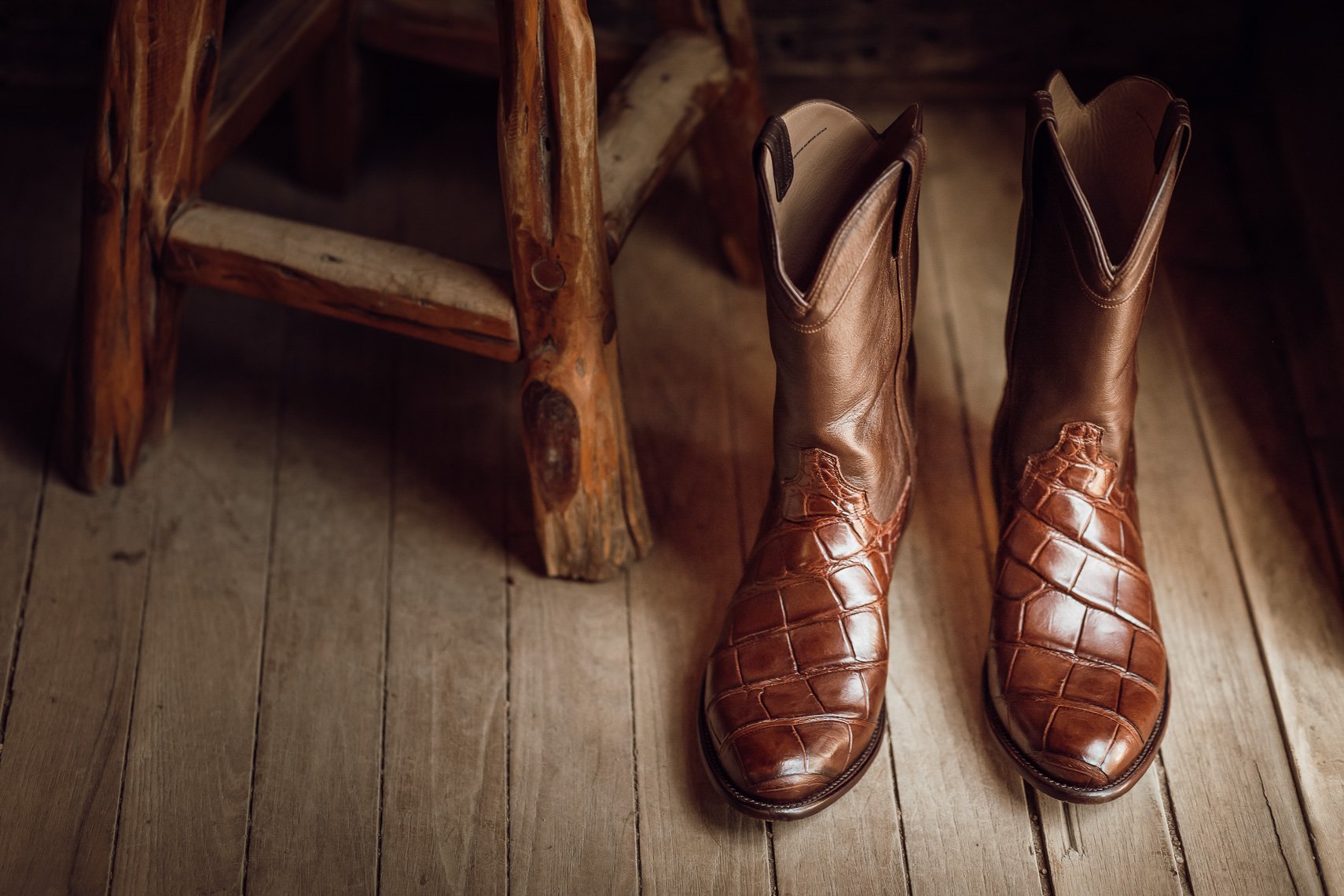 A pair of brown cowboy boots with crocodile pattern detail on the toe and foot area, placed on a wooden floor next to a rustic wooden chair.
