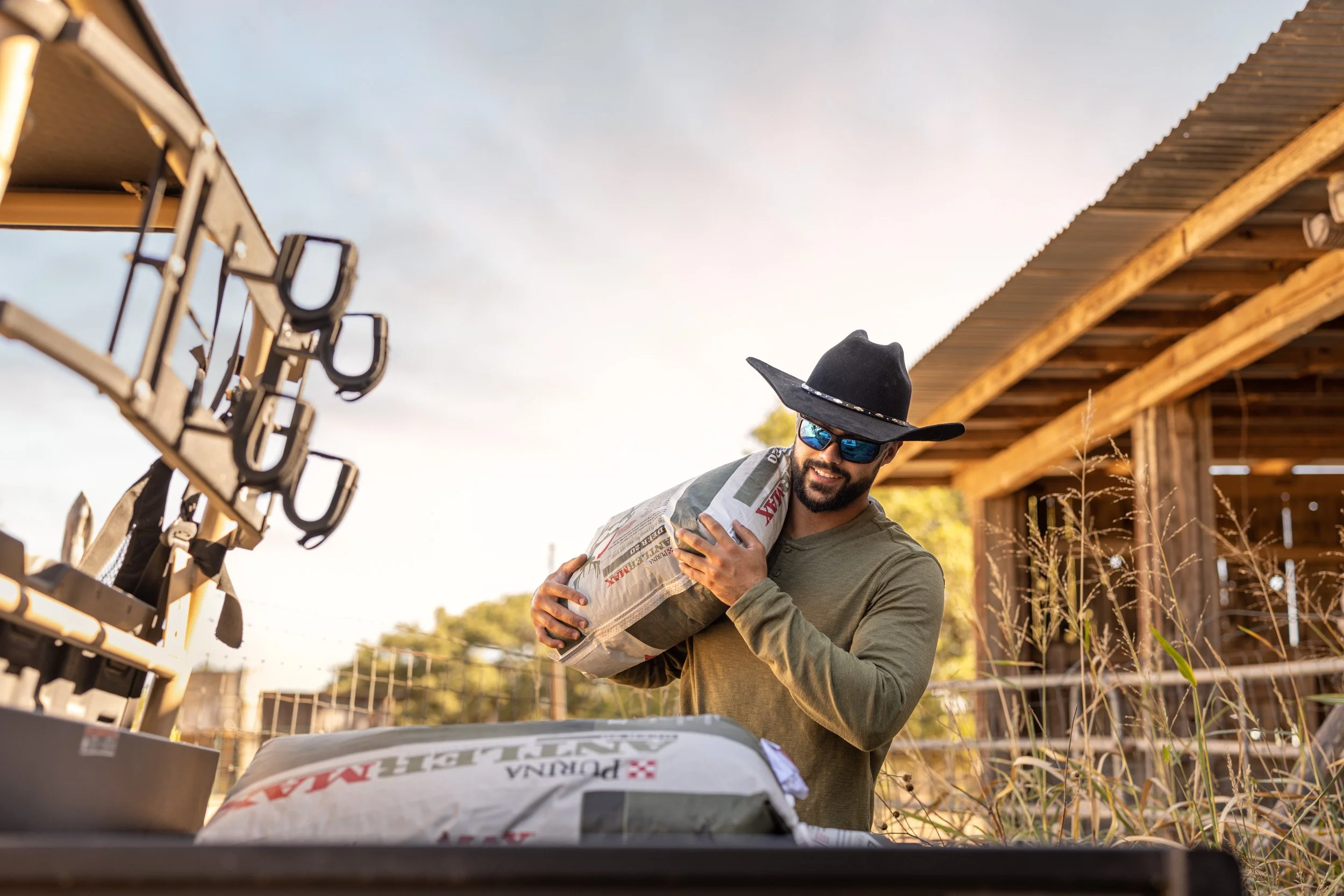 Man wearing sunglasses and a wide-brimmed hat holds a bag of fertilizer or seed while standing outdoors near a wooden structure, with equipment and tools visible on a table in the foreground.