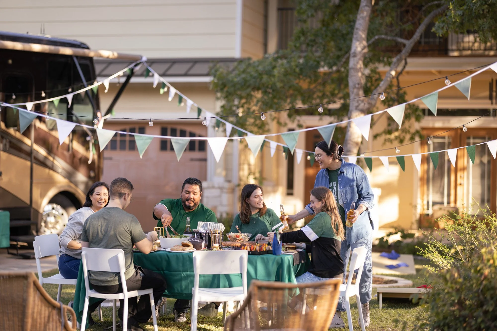 People enjoying an outdoor gathering with a table of food and drinks, under string lights and banners in a residential backyard.