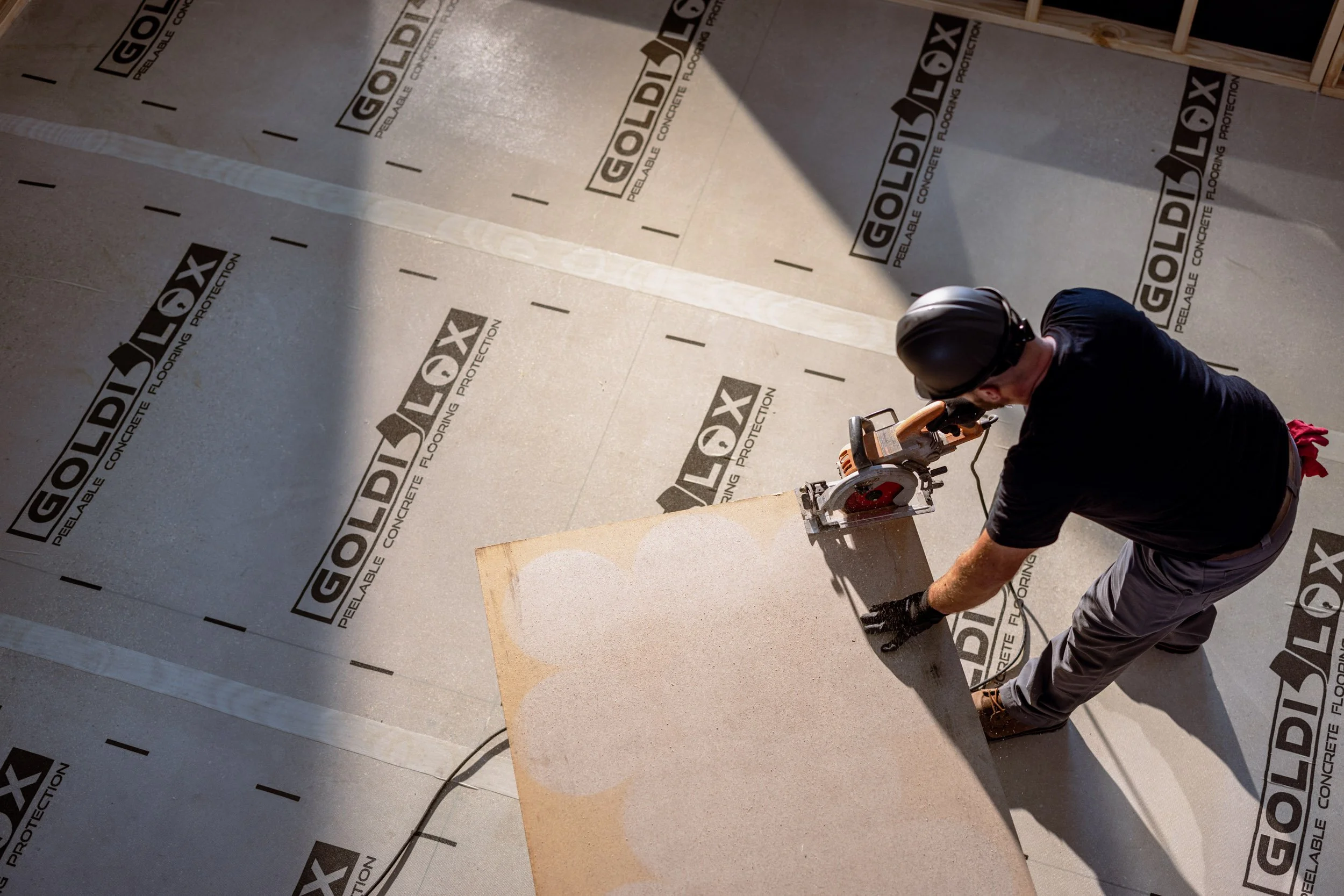 A worker in a black shirt and gray pants using a power saw on a wooden board on a floor covered with protective material that has the brand name GOLIDEX printed repeatedly.
