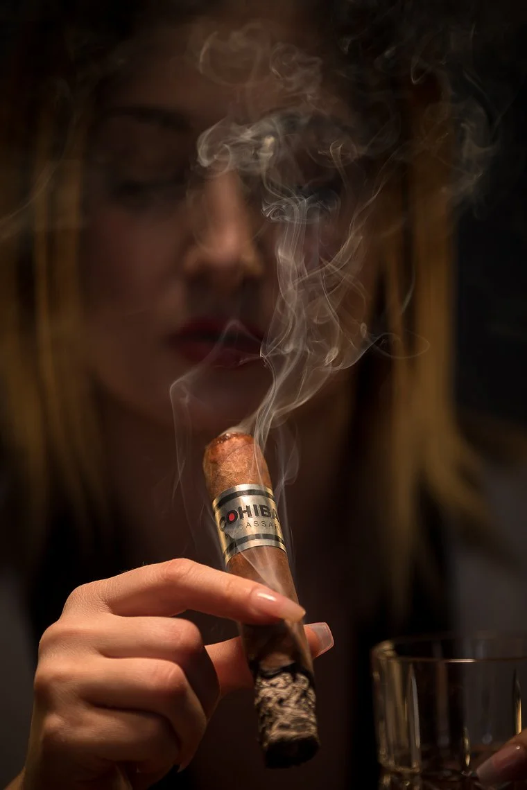 A woman with blonde hair and red lipstick holding a lit cigar in her hand, smoke rising from the cigar, and a glass in front of her.
