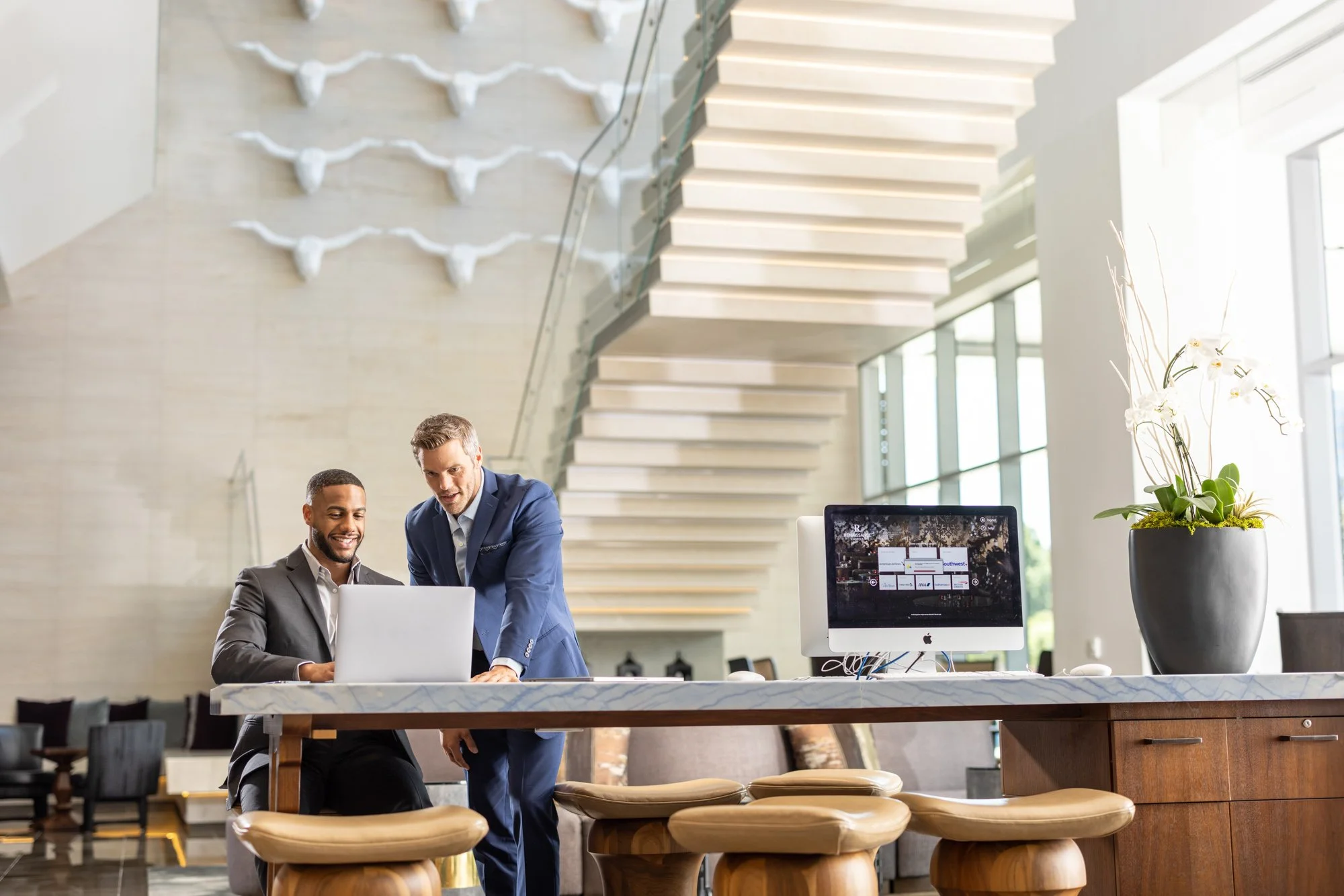 Two men in business attire working together at a computer in a modern, bright office with large windows, stairs in the background, and decorative wall art featuring animal head sculptures.