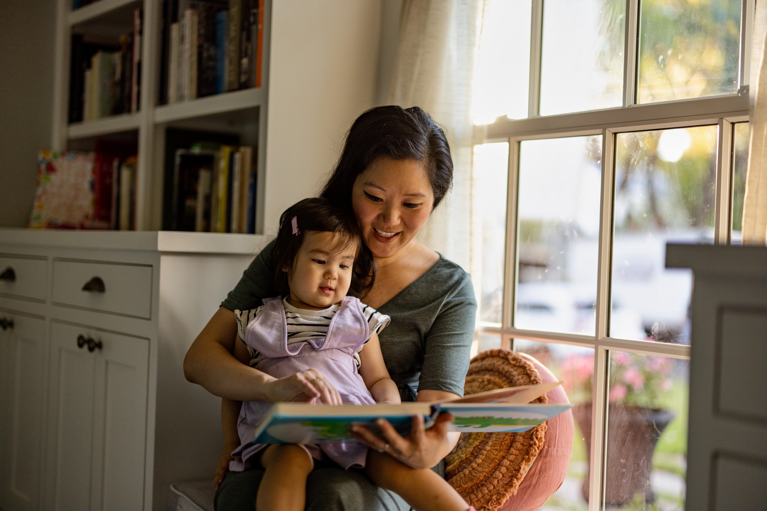 A woman sitting by a large window, holding a young girl while looking at a colorful children's book. The room has bookshelves and a white dresser, and the sunlight is streaming through the window.
