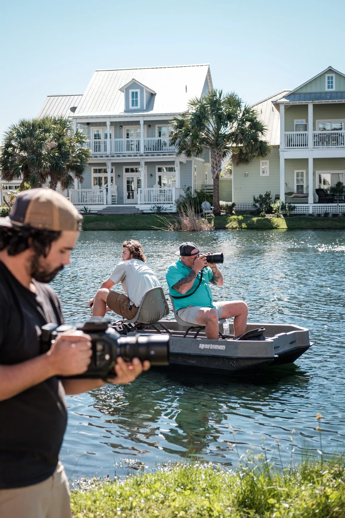Two men on a boat in a waterway, one taking photographs, and another looking at his camera. Two other men are in the foreground, one holding a camera or phone. Background features a large white house with balconies, palm trees, and a clear blue sky.