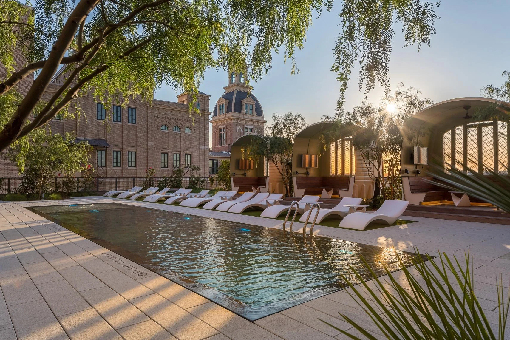 A luxurious outdoor pool area at sunset with modern white lounge chairs and curved cabanas, surrounded by lush green trees and a historic brick building with a domed tower in the background.