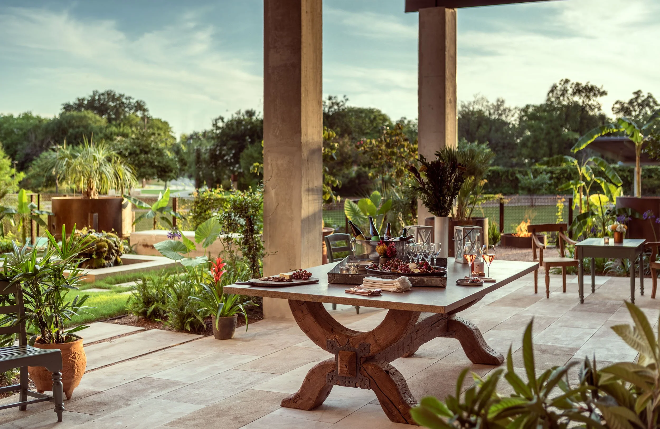 Outdoor patio with a wooden dining table set for a gathering, surrounded by lush green plants, trees, and a fire pit in the background, under a partly cloudy sky.