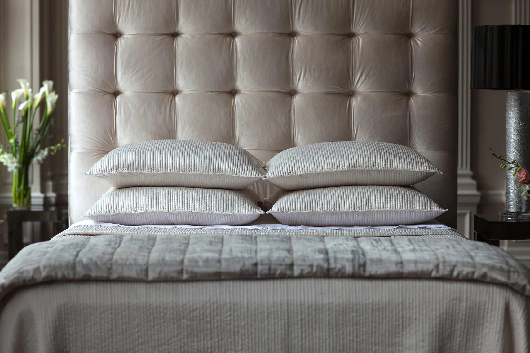A neatly made bed with a beige, tufted upholstered headboard, four white striped pillows stacked two by two, and a patterned bedspread in muted tones. On either side of the bed are bedside tables, with a vase of white flowers on the left and a gray l