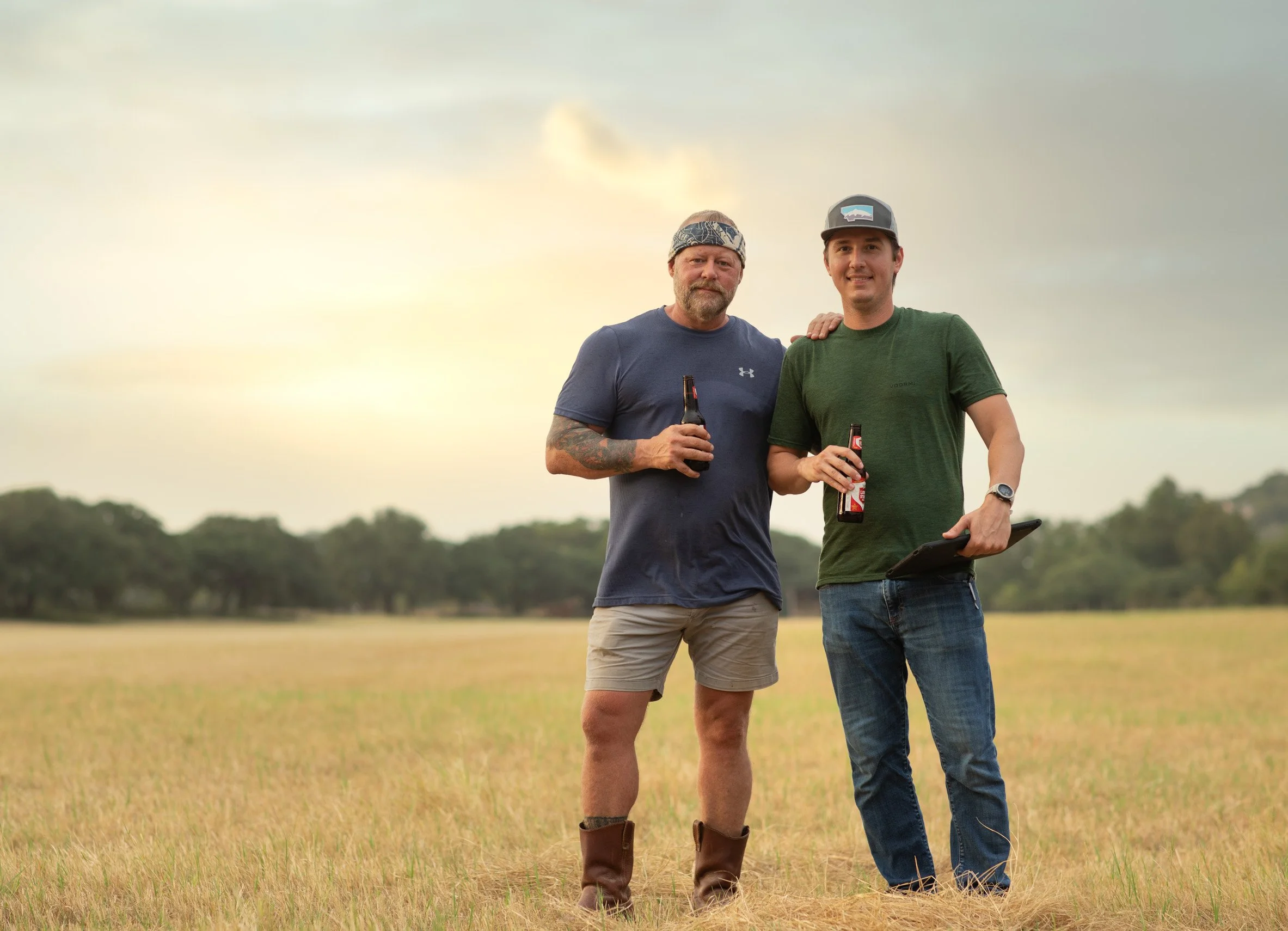 Two men standing in a grassy field at sunset, holding beer bottles, with one man resting his hand on the other's shoulder, smiling.