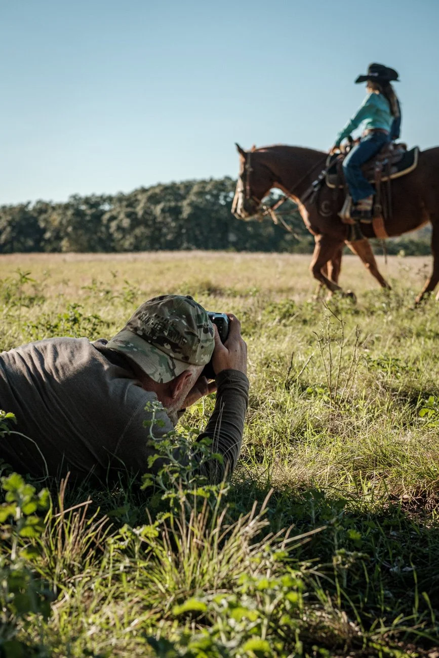 A person in camouflage clothing lying on the grass taking a photo of a woman on horseback riding across a grassy field on a clear, sunny day.