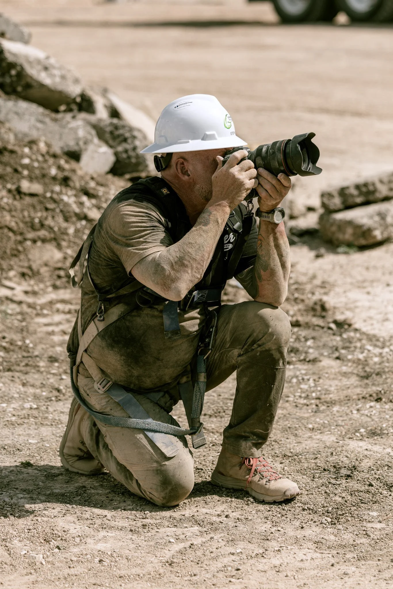 A man kneeling on one knee on a rocky, dusty construction site during the day, taking a photograph with a professional camera, wearing a white hard hat, sunglasses, a tan shirt, and beige pants with a harness and work boots.