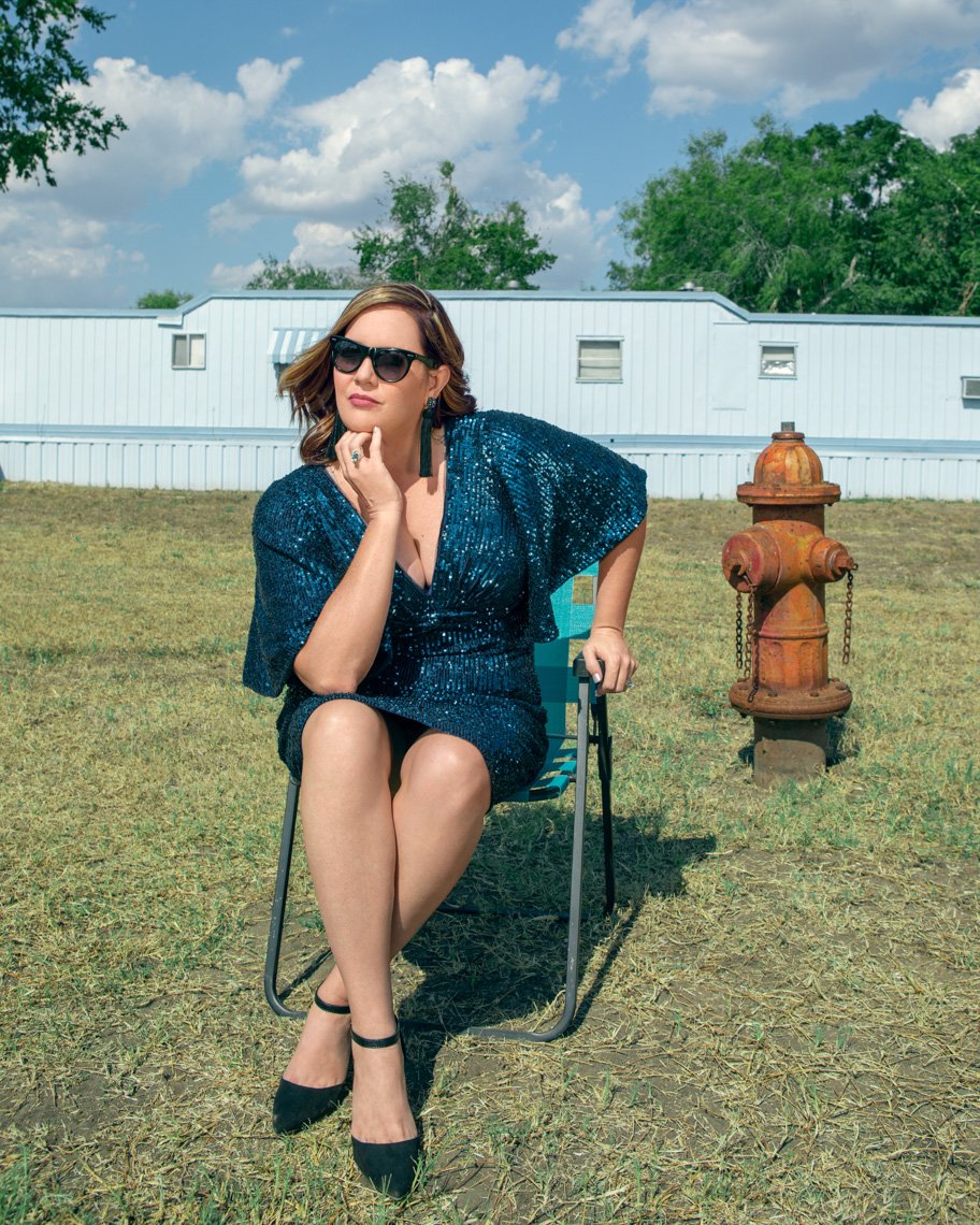 A woman with short brown hair, wearing sunglasses, a dark blue sequin dress, and black shoes, is sitting on a folding chair outdoors on grass with a rusty fire hydrant nearby. She is resting her chin on her left hand, with a contemplative expression,