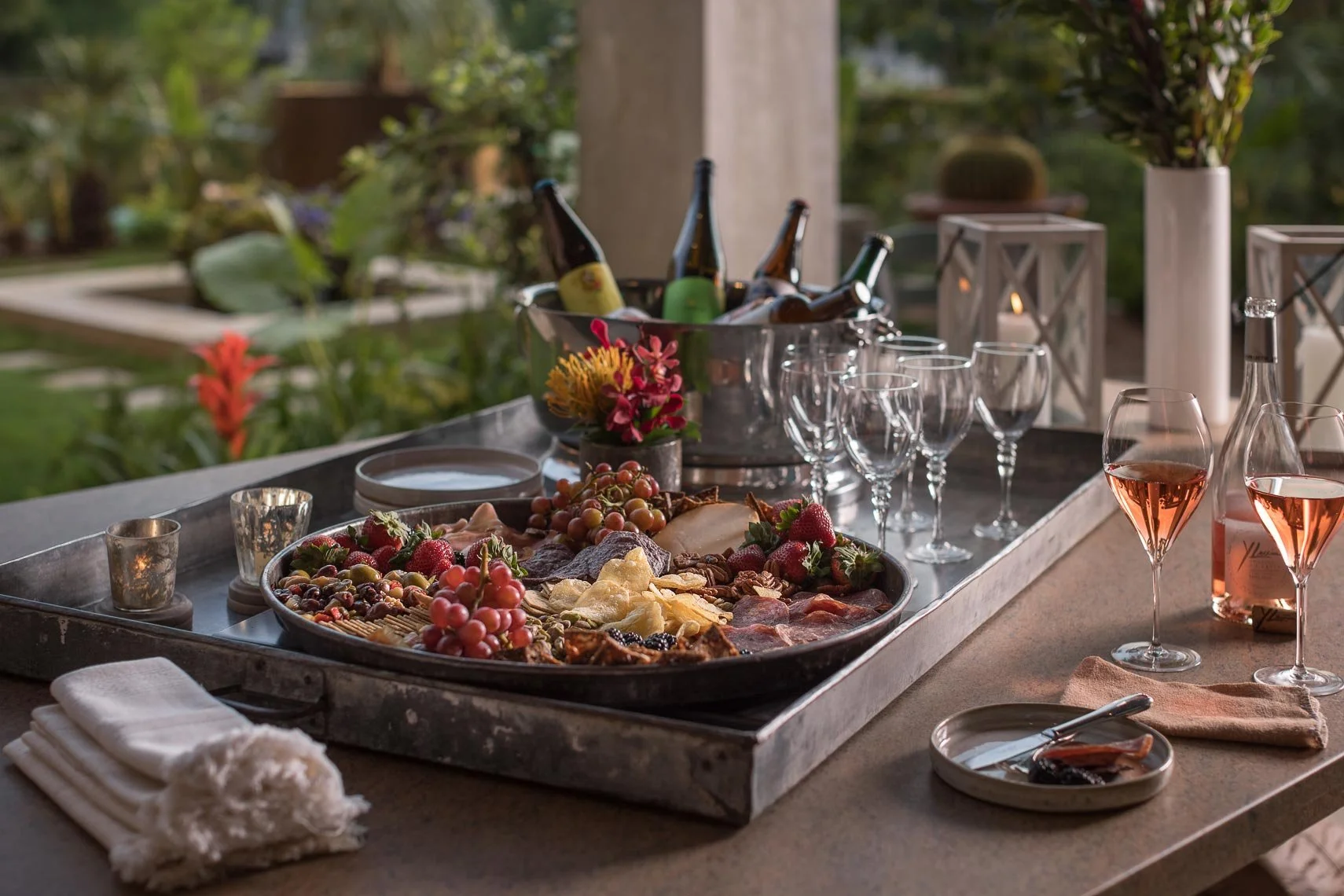 Outdoor dinner table with a charcuterie board of cheese, grapes, berries, and meats, wine glasses filled with rosé, and a large bowl of beer bottles in an outdoor garden setting.