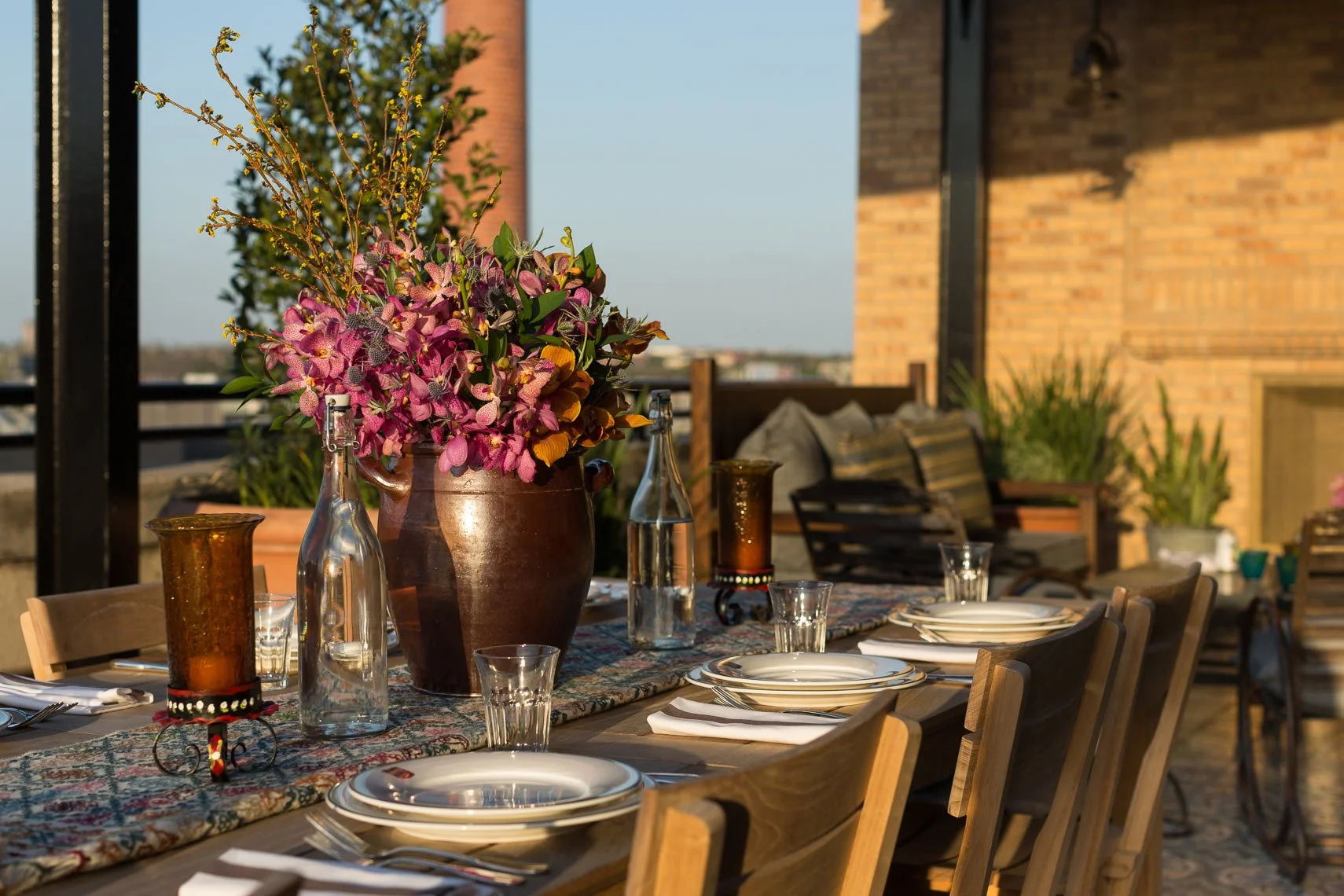 An outdoor dining table set for a meal with a large flower arrangement in a brown vase, glass bottles, and candles, with chairs around it on a balcony or terrace with a cityscape view and a brick wall in the background.