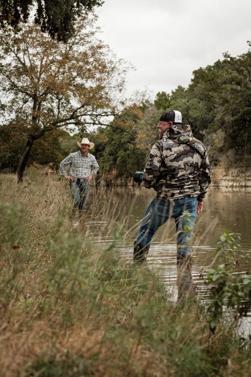Two men by a riverbank surrounded by trees, one in camouflage jacket and cap holding a camera, the other in cowboy hat and checkered shirt smiling.