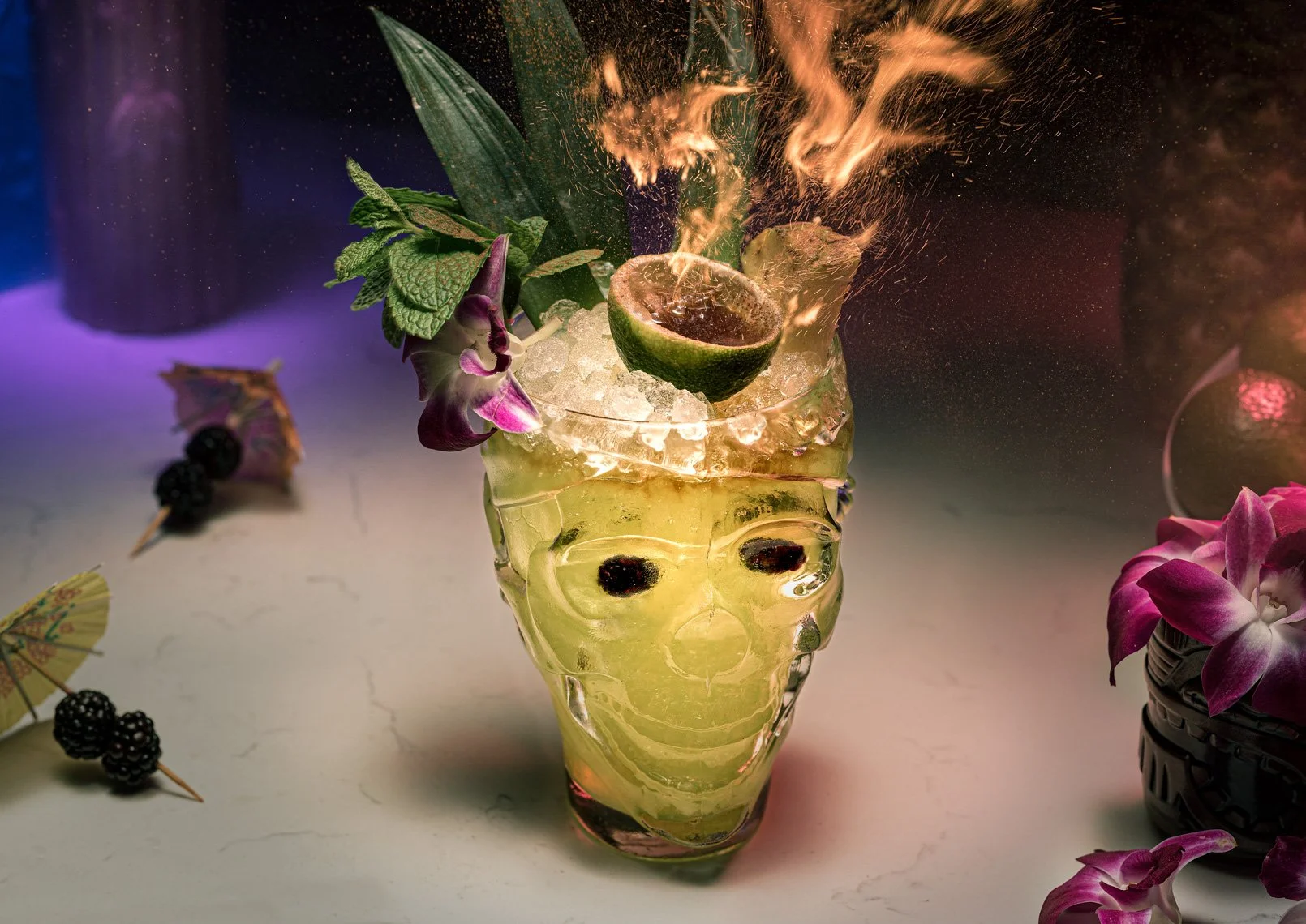 A tiki-style glass filled with a green cocktail, garnished with mint, flower, blueberries, and ice, with flames and sparks exploding from a lime on top, surrounded by flowers and decorative skewers.