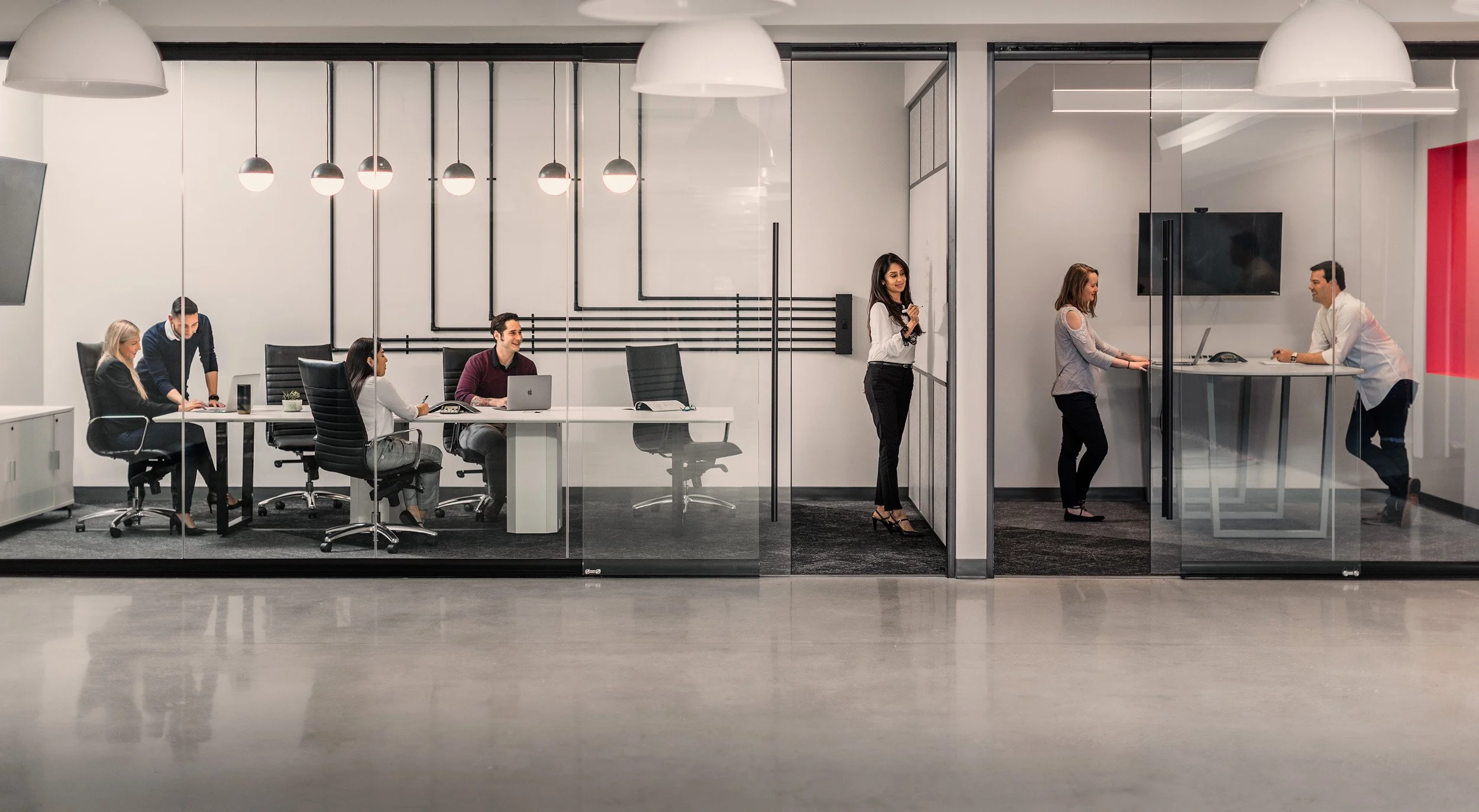 A modern office conference room with five people engaged in a meeting, viewed through a glass wall. Four individuals are seated at a white table working on laptops, while one woman stands by in front of a digital TV screen and appears to be speaking 