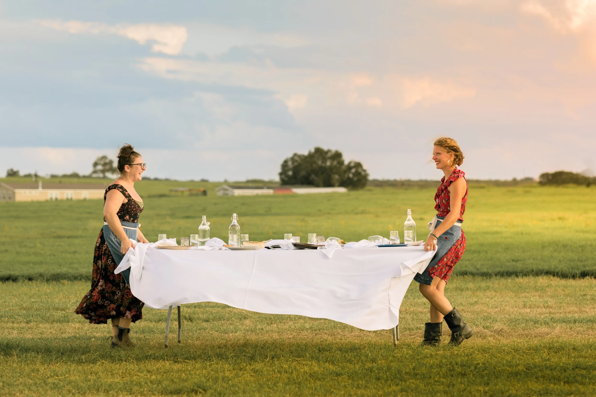 Two women in dresses and boots setting up a long outdoor table with white tablecloths and bottles in a green field with trees and farm buildings in the background, during sunset.