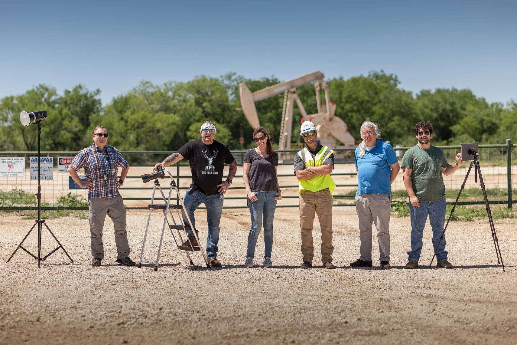 Group of six people standing outdoors in front of a fence and an oil pumpjack, with equipment like cameras and a tablet on tripods, under clear blue sky.