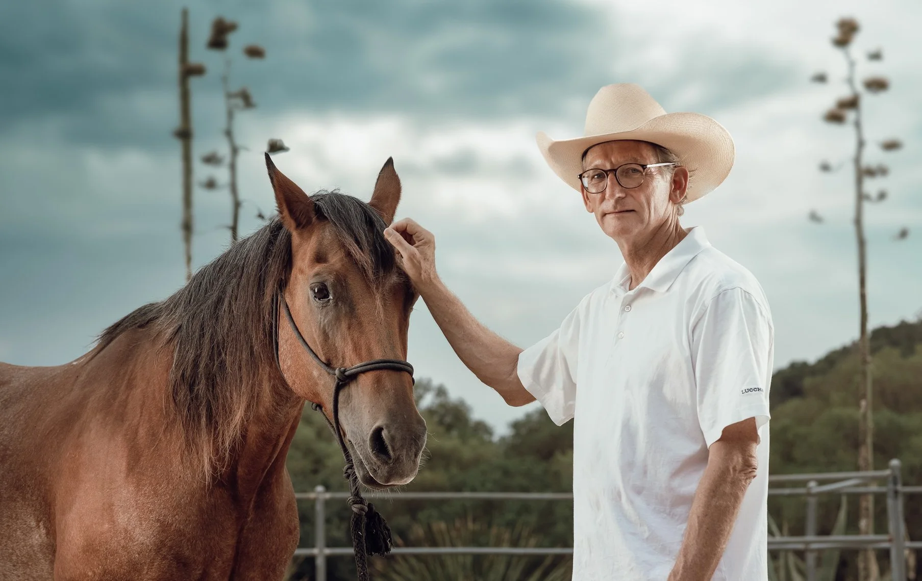 An elderly man wearing a white cowboy hat, glasses, and a white polo shirt stands next to a brown horse with a black mane. The man is gently touching the horse's forehead. In the background, there are tall trees, overcast skies, and a distant fence.