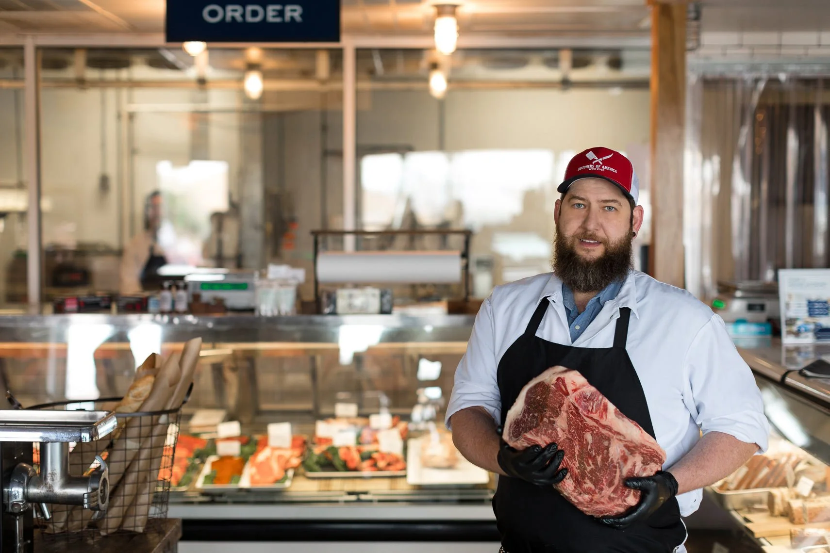 A man with a beard wearing a red baseball cap, a white shirt, and an apron holding a large piece of raw steak inside a butcher shop or meat market.