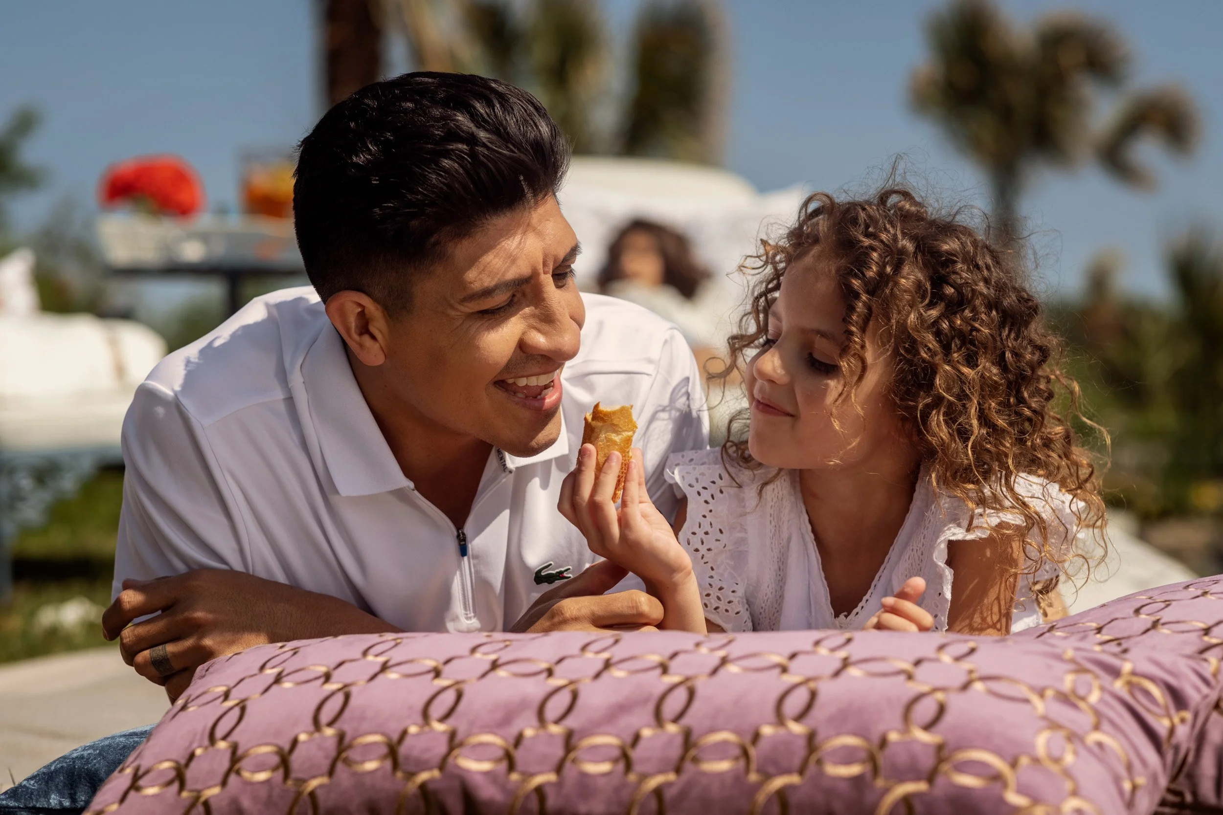 A man and young girl sharing a bite of food outdoors on a sunny day, with a table and people in the background.