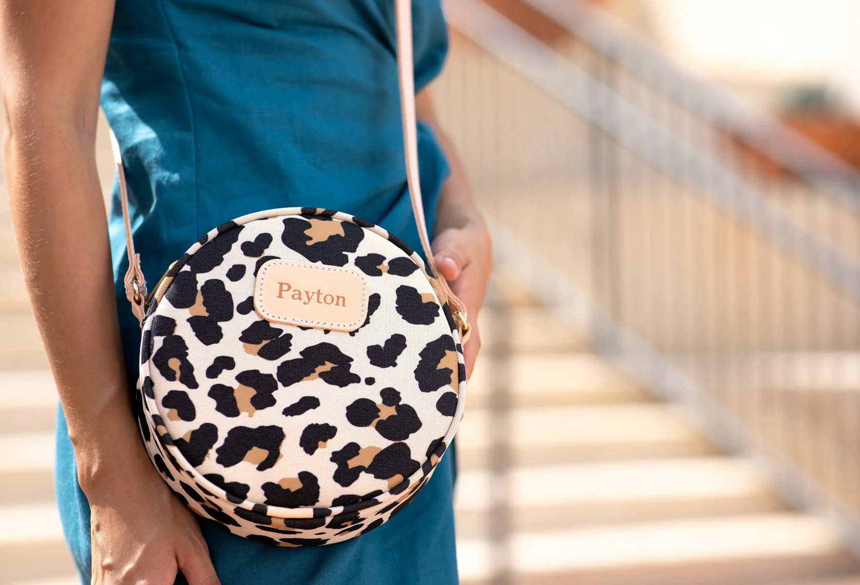 Person holding a small round leopard-print bag with a pink strap and a name tag that reads Payton, standing on a staircase with sunlight in the background.