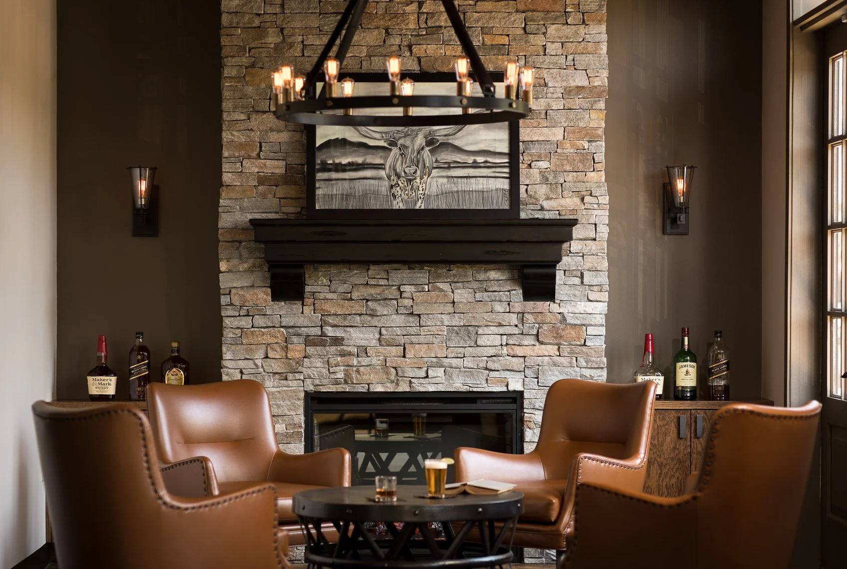 A cozy living room with a stone fireplace, a large framed black and white painting of a deer, brown leather chairs, a round black coffee table with drinks, and a chandelier hanging from the ceiling. There are bottles of liquor on a side cabinet and w