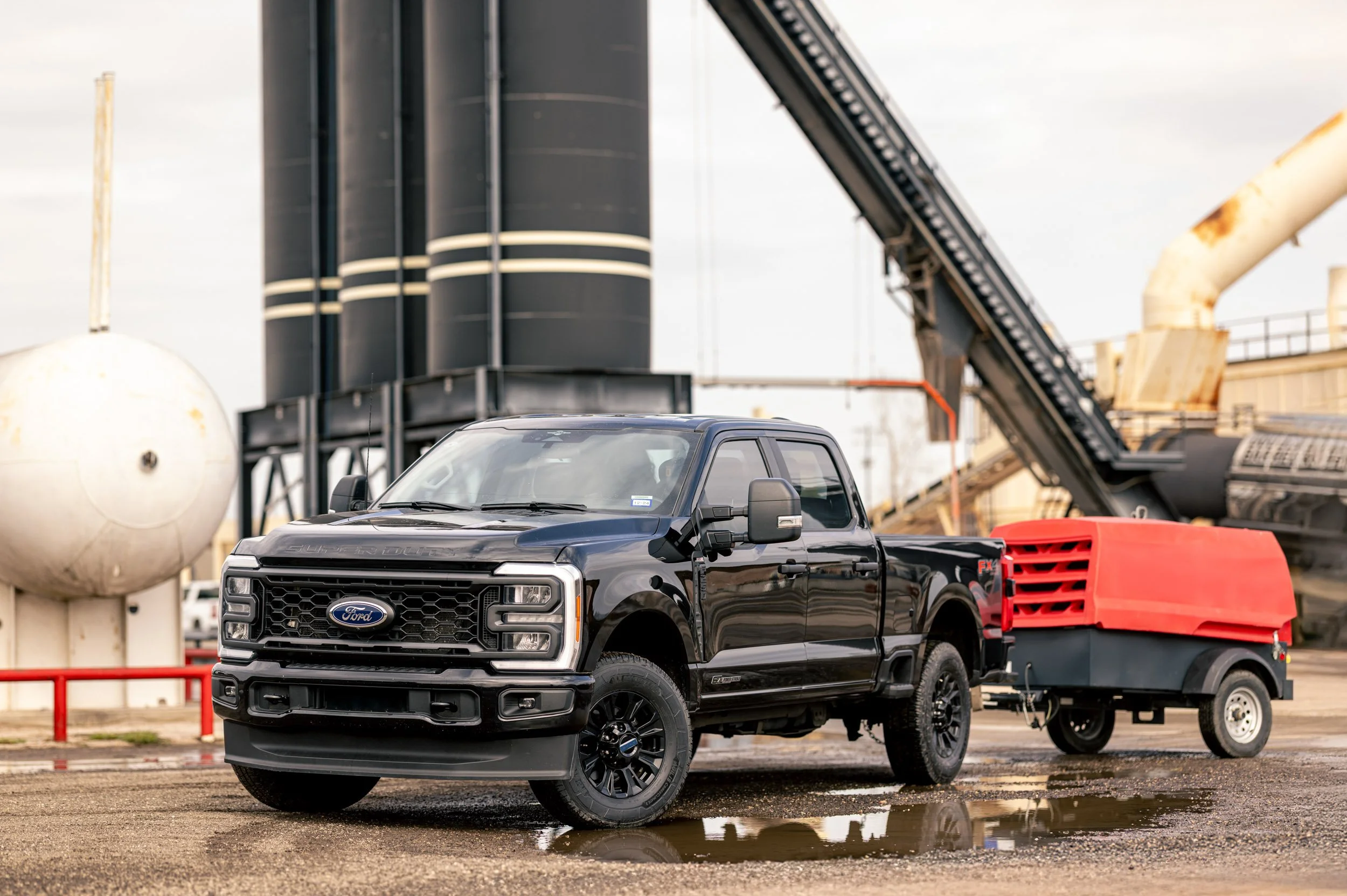 A black Ford pickup truck parked in an industrial lot next to a trailer with a red container. In the background, there are large industrial tanks and a conveyor belt structure.