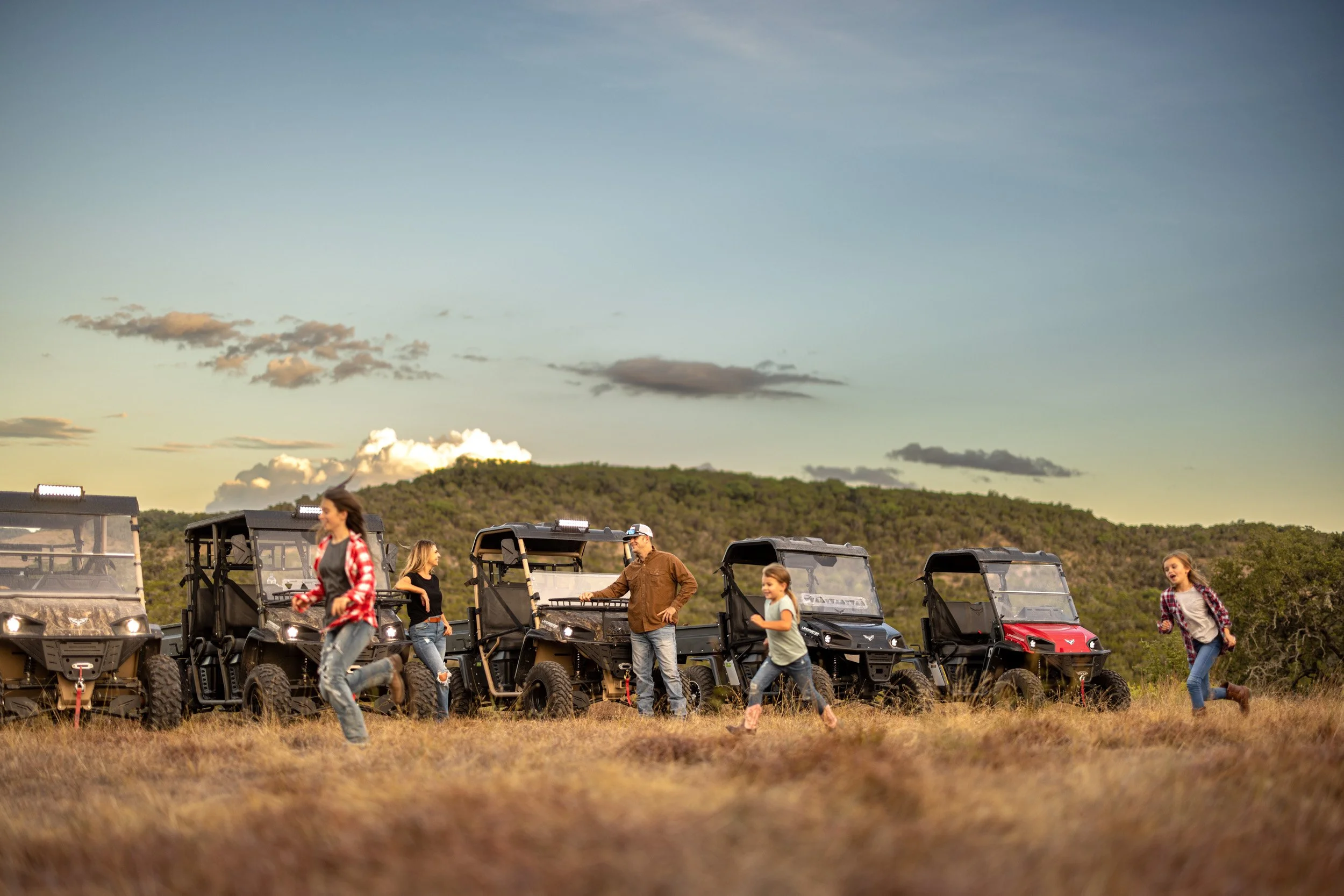 People outdoors near black utility vehicles on a grassy field with hills and a partly cloudy sky in the background.