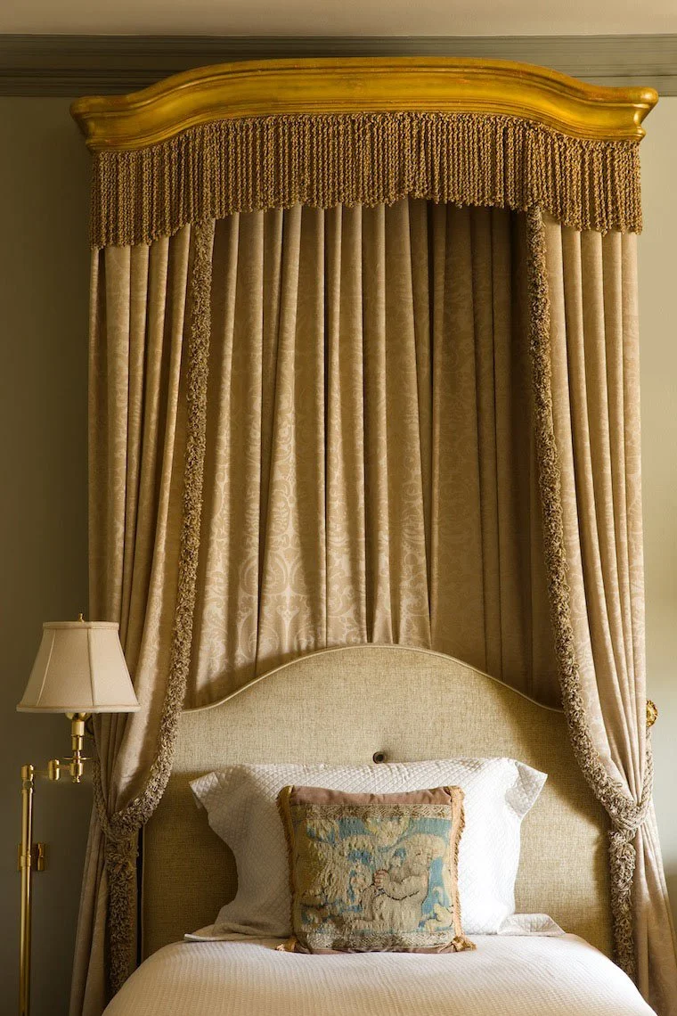 A bed with a tall, ornate beige headboard and a beige canopy with decorative trim. There is a white pillow and a small decorative pillow with a tapestry design on the bed. A gold-colored bedside lamp with a white shade is on the left side.