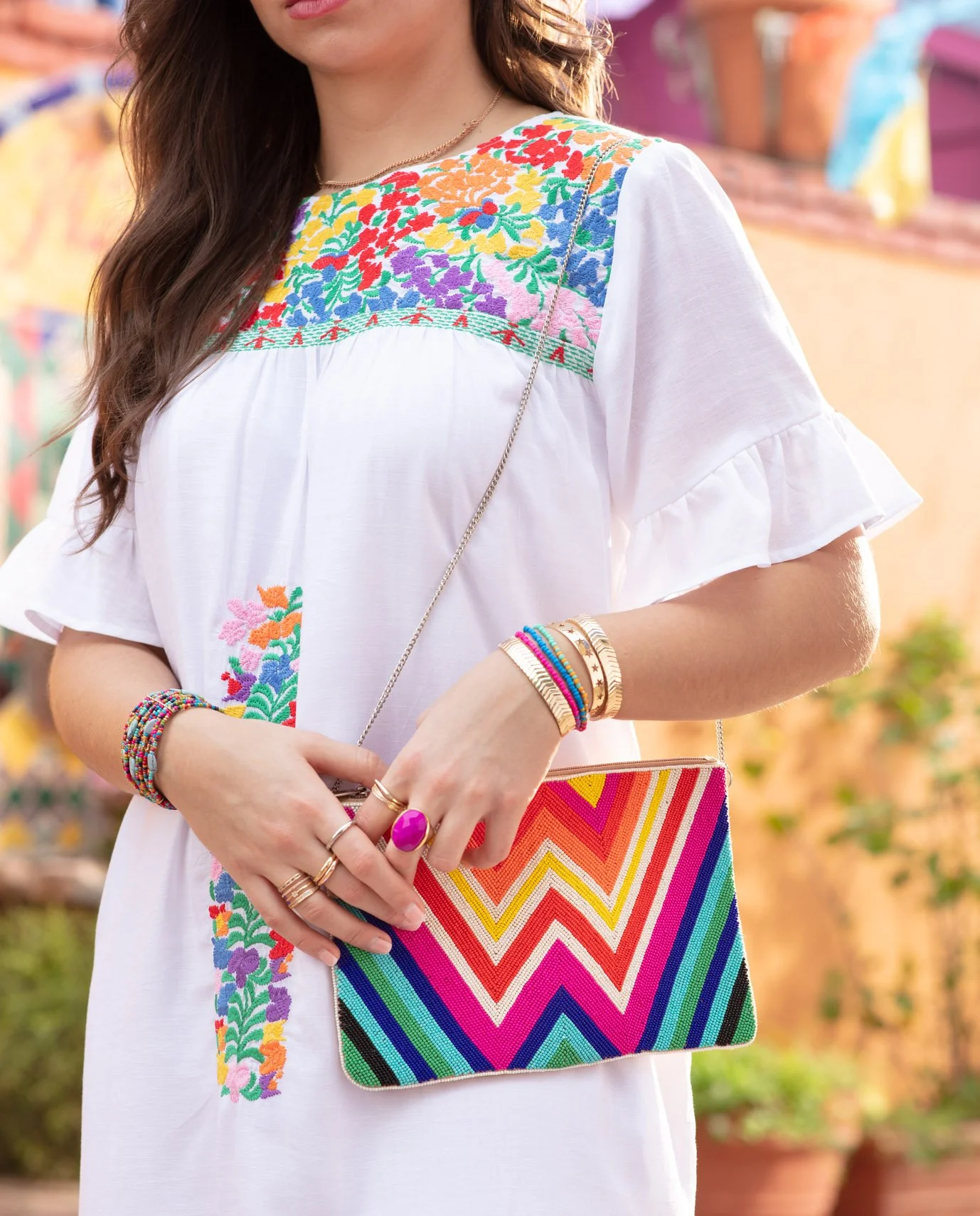 A woman wearing a white embroidered dress with colorful floral patterns, holding a rainbow-colored zigzag patterned clutch, wearing jewelry, and standing outdoors with blurred colorful background