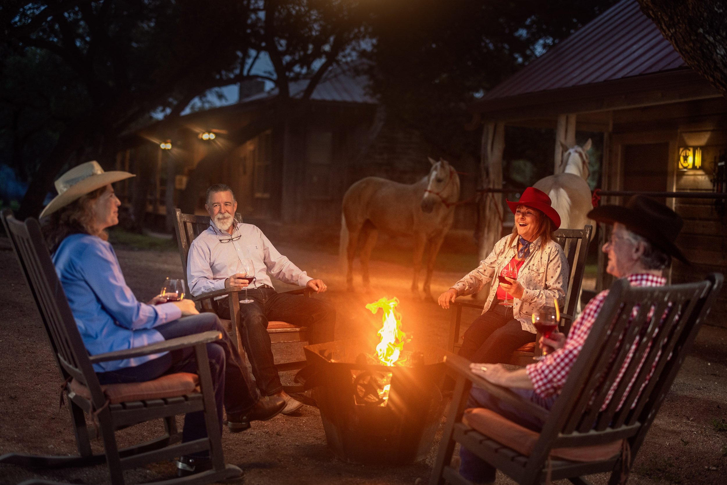 A group of five older adults sitting around a campfire in a backyard at night, with two horses and a wooden house in the background. They are dressed in casual and western-style clothing, some with hats, and are enjoying drinks and conversation.