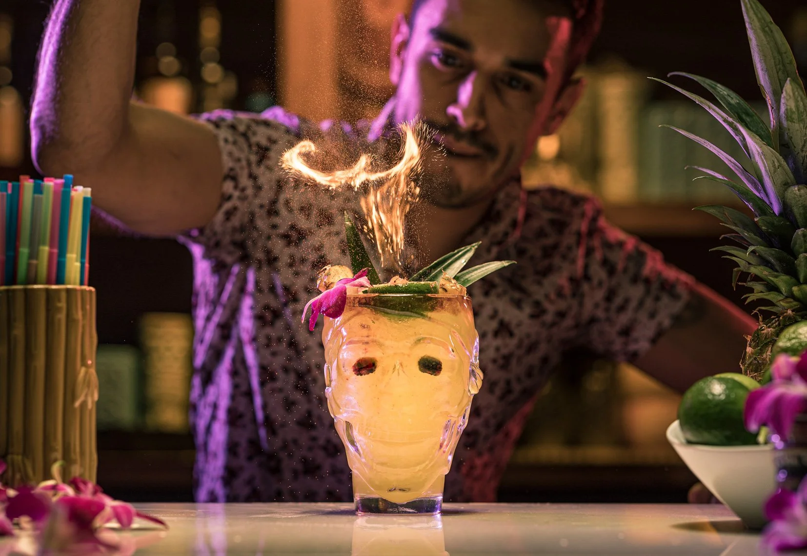 A bartender making a flaming tropical cocktail in a pineapple-shaped glass, garnished with pineapple leaves and pink flowers, with colorful straws on the side and a pineapple on the right.