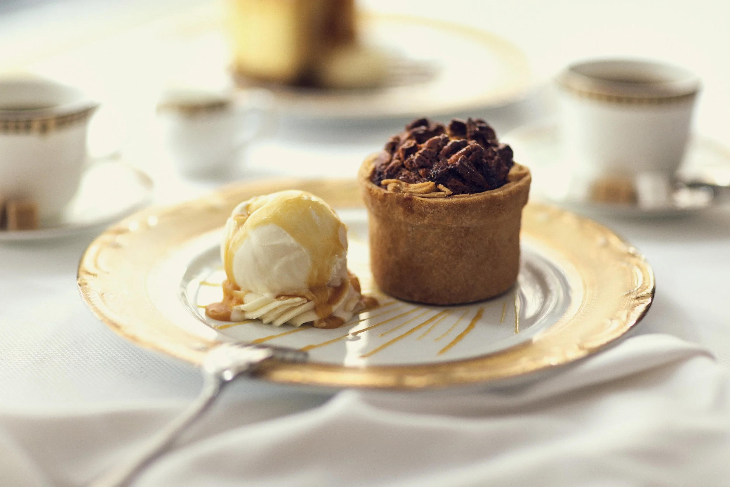 Three assorted mini desserts on a decorative plate, including a chocolate-topped tart, a white chocolate mousse, and a cream-filled pastry, with a blurred background of teacups and plates.