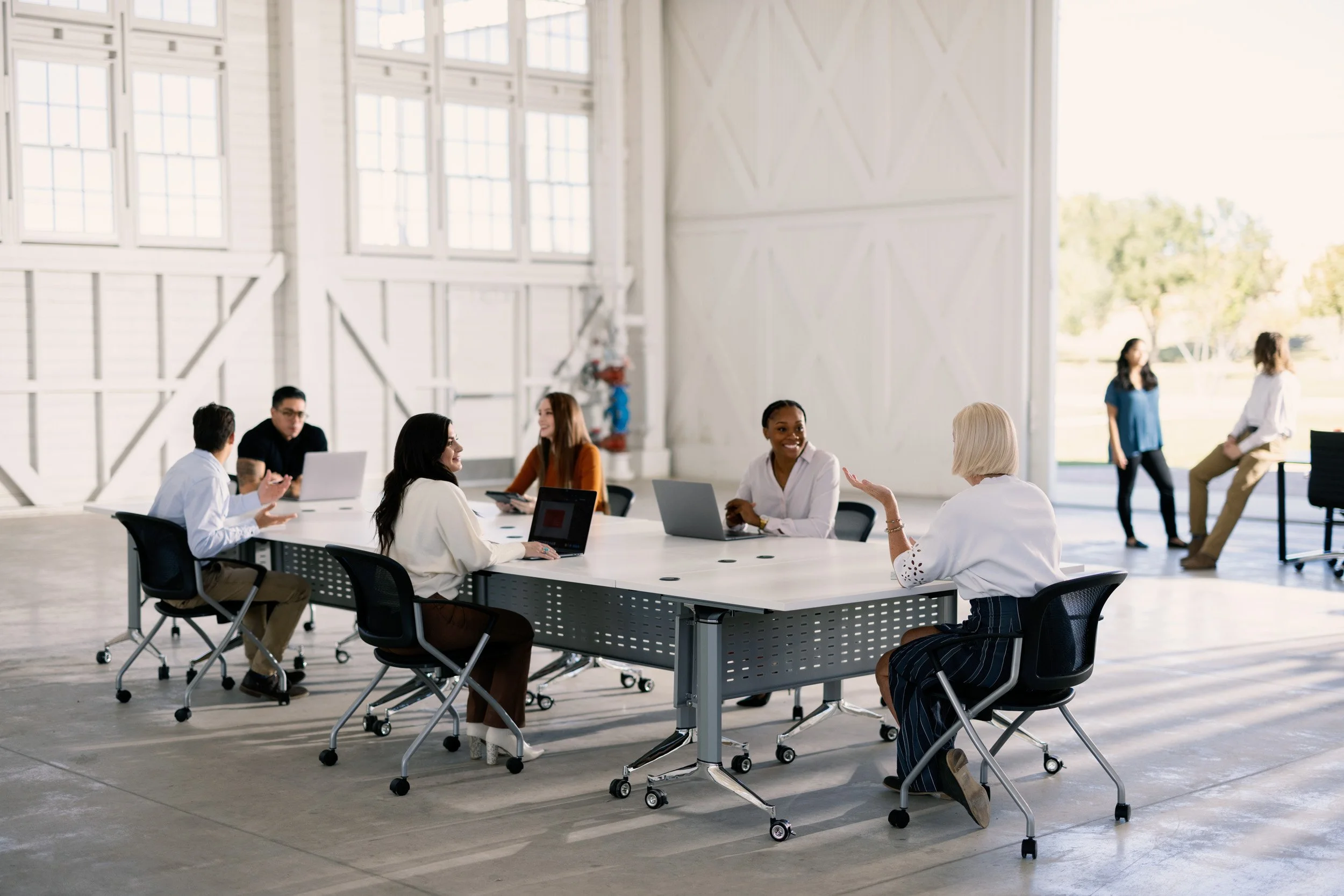 A group of diverse people engaged in a discussion around a large white table in a bright, modern meeting room with large windows and white walls.