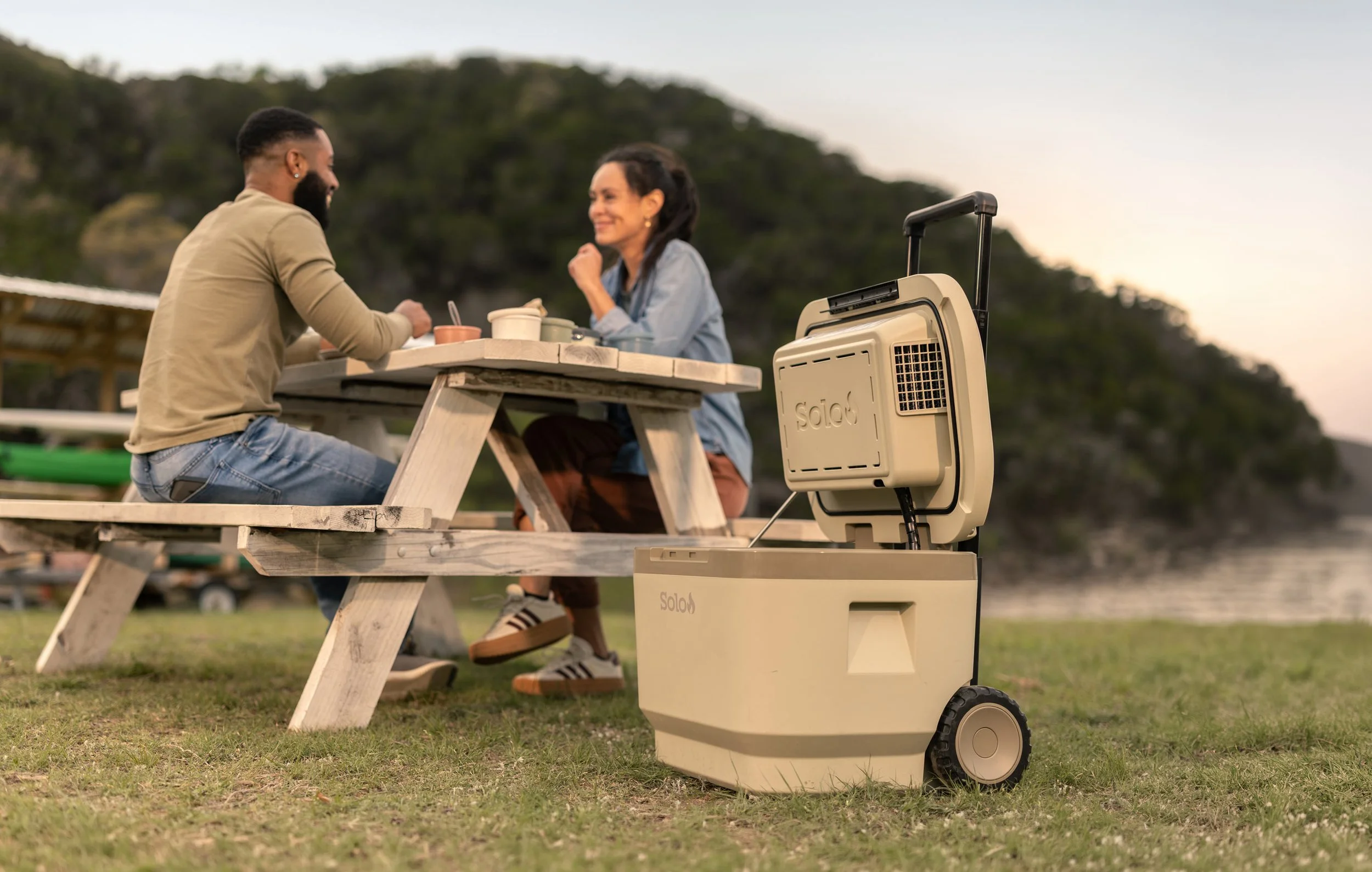 A man and a woman are sitting at a picnic table outdoors, smiling and enjoying a conversation. There is a portable cooler on the ground next to them, with a device on top of it. The background shows green hills and a clear sky, indicating a pleasant 